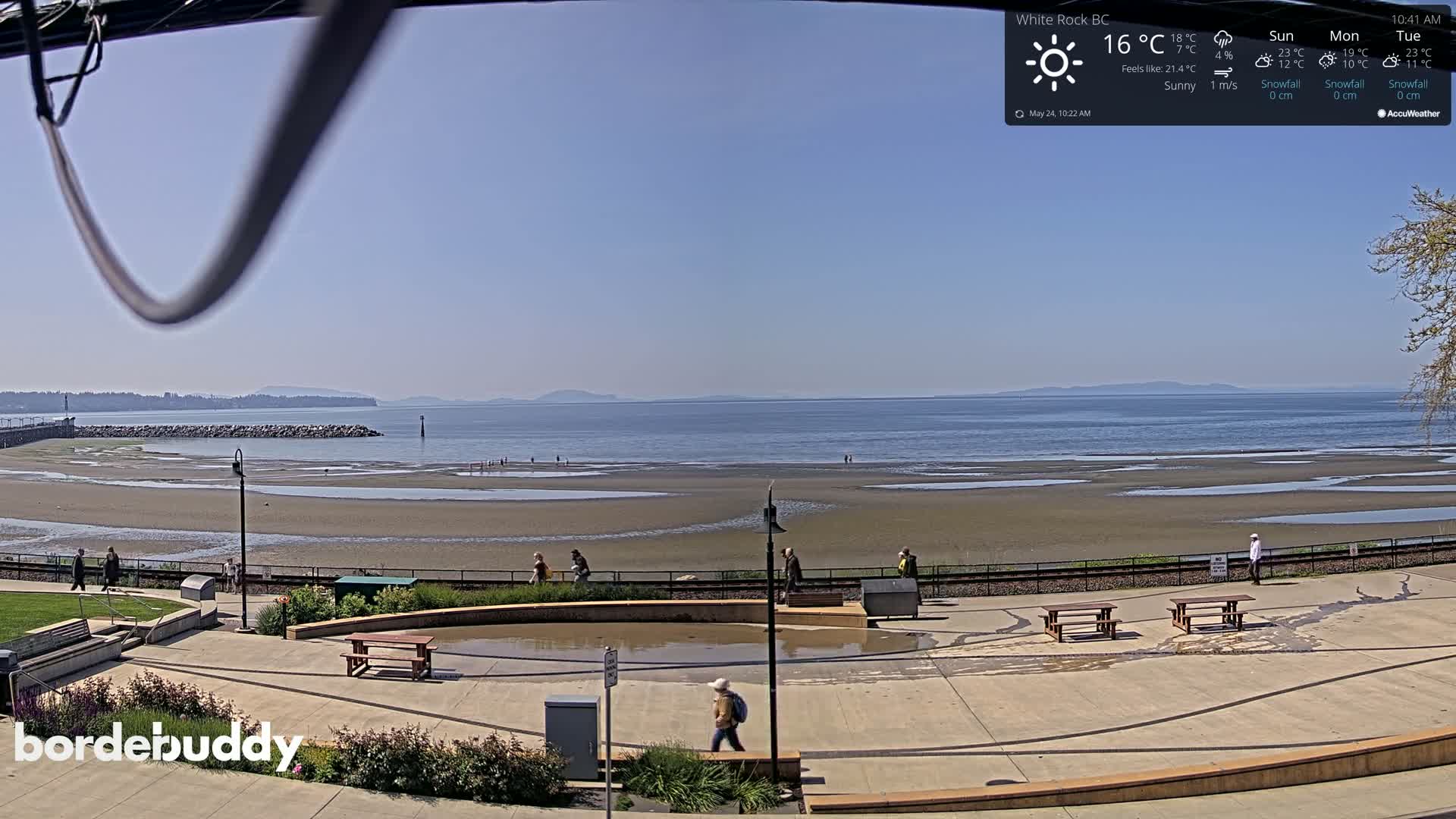 A sunny beach with a wide expanse of exposed sand and calm water, showing people walking along a paved walkway near picnic tables and a water feature.
