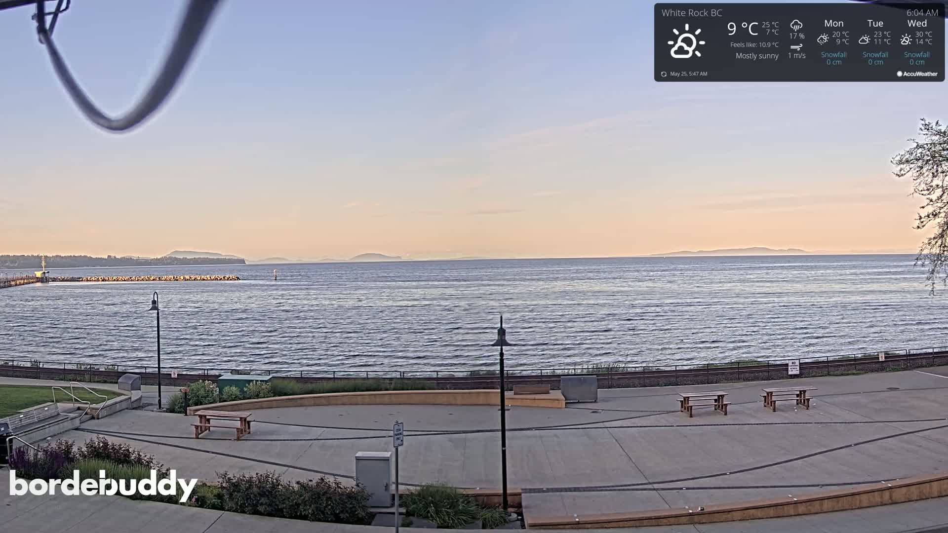 A mostly sunny, slightly cool day overlooking a calm ocean with distant mountains and a paved waterfront area with benches.