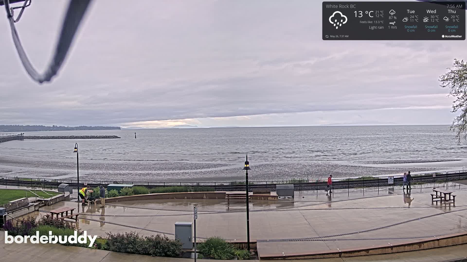 A wet, paved waterfront area overlooks a calm ocean under a cloudy sky with light rain.