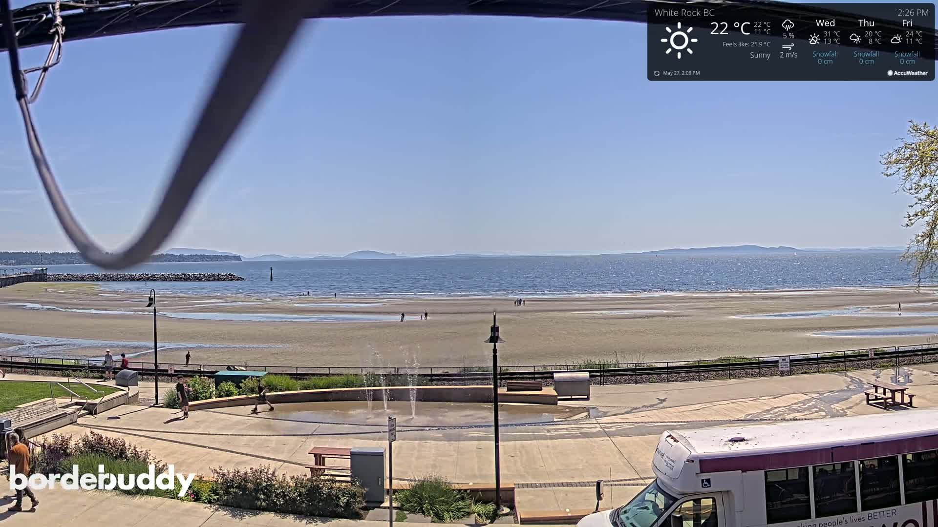 A sandy beach with a small fountain and a few people is visible, with a calm ocean and distant mountains under a sunny sky.