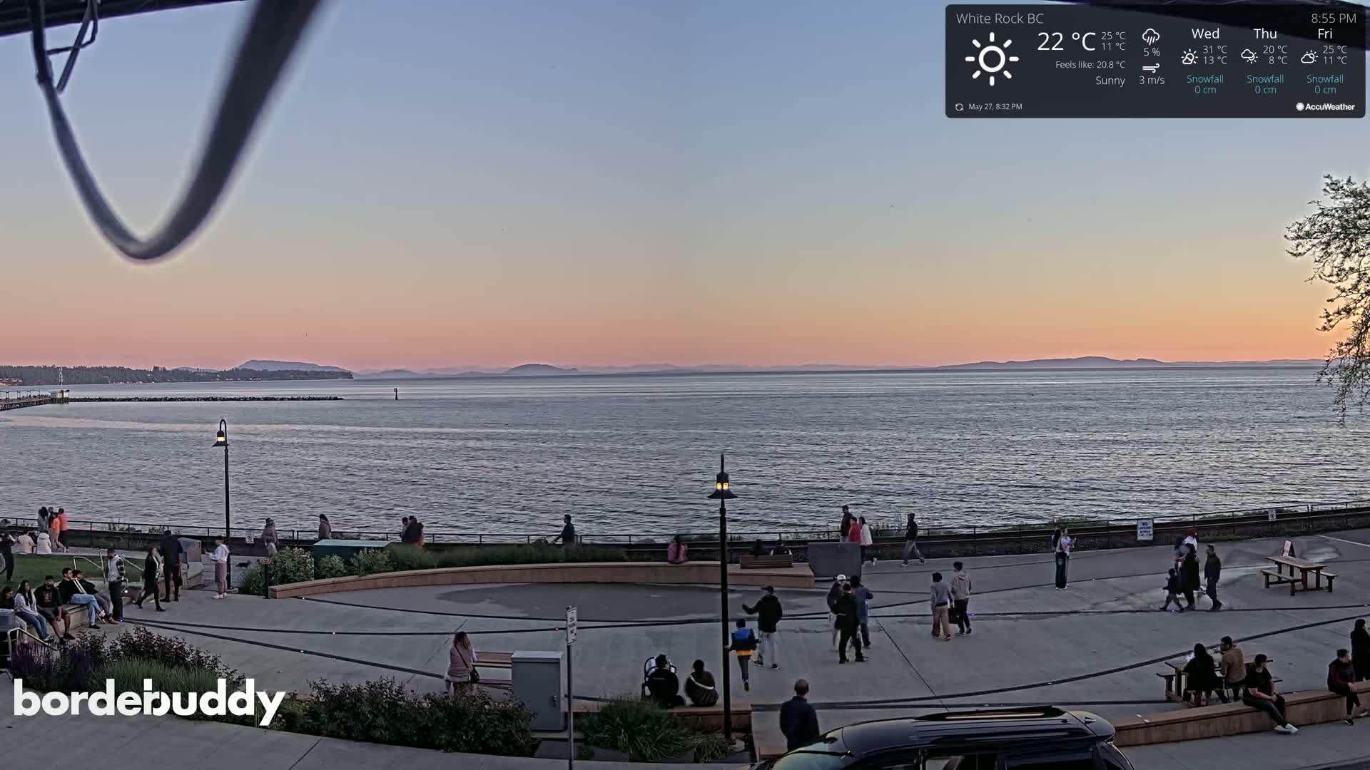 A calm body of water at sunset, with people gathered on a paved waterfront area under a clear, sunny sky.
