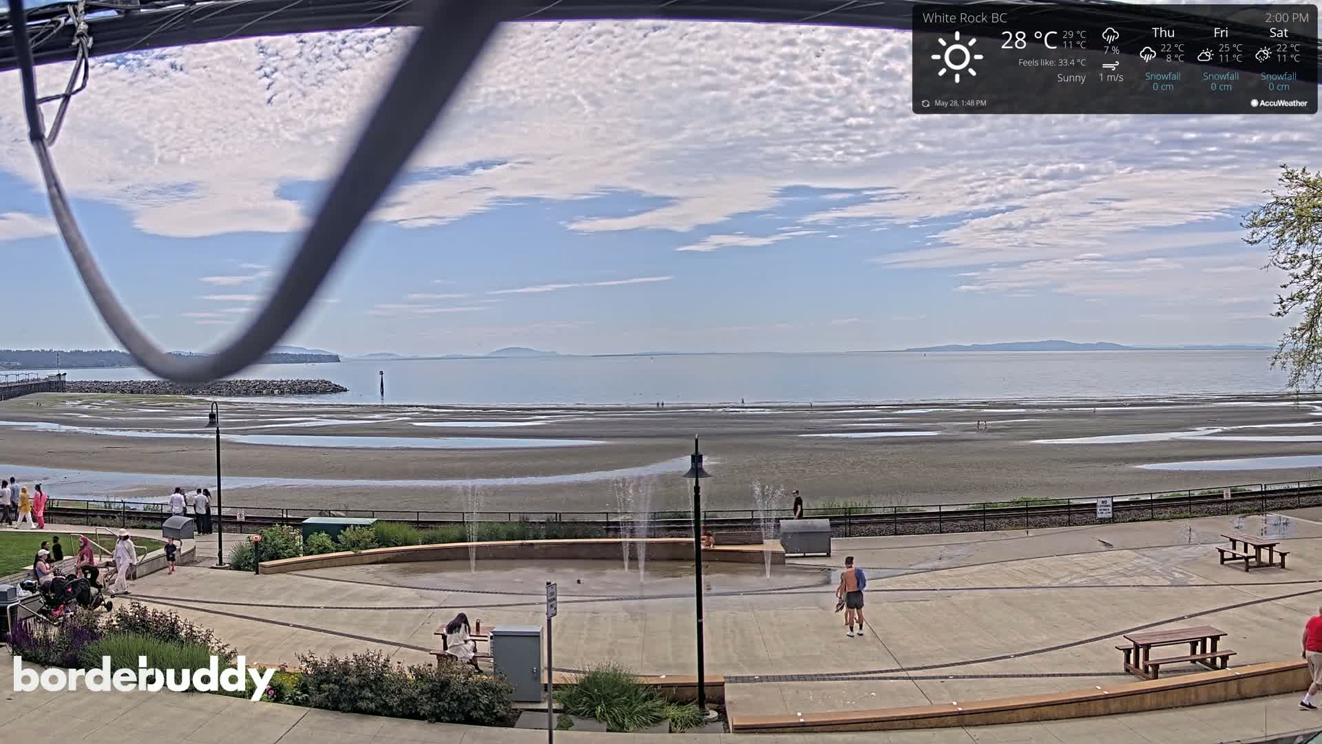 A sunny beach scene with a calm ocean, a paved area with small water features, and several people enjoying the day.