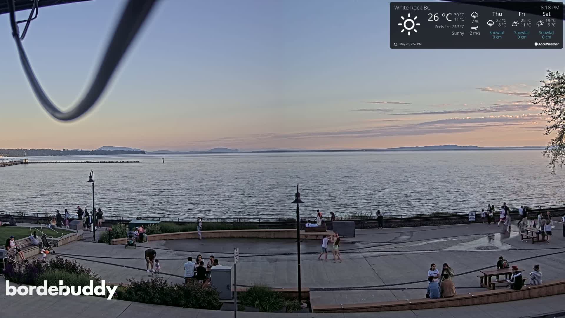 A sunny evening scene overlooks a calm body of water with people gathered along a waterfront promenade.