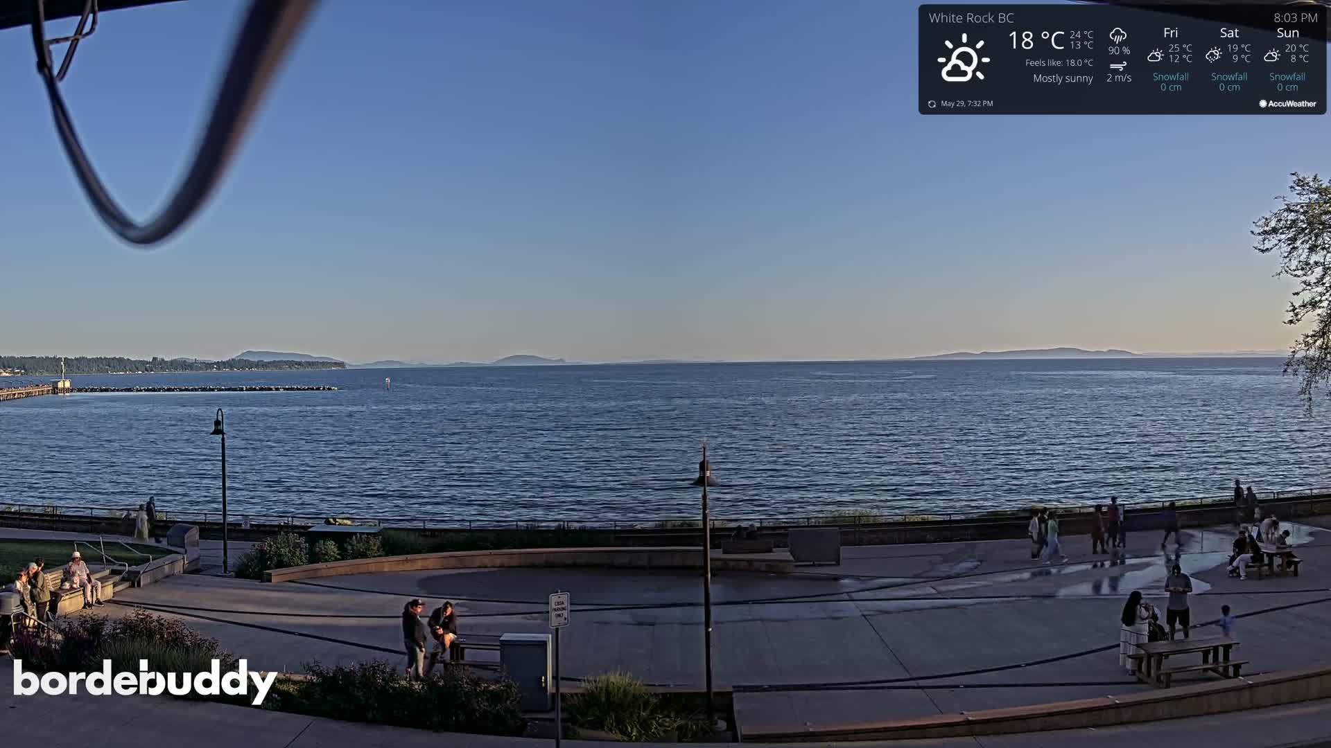 A mostly sunny waterfront scene shows a calm body of water with distant mountains under a clear blue sky, featuring people relaxing on a paved area near the water's edge.