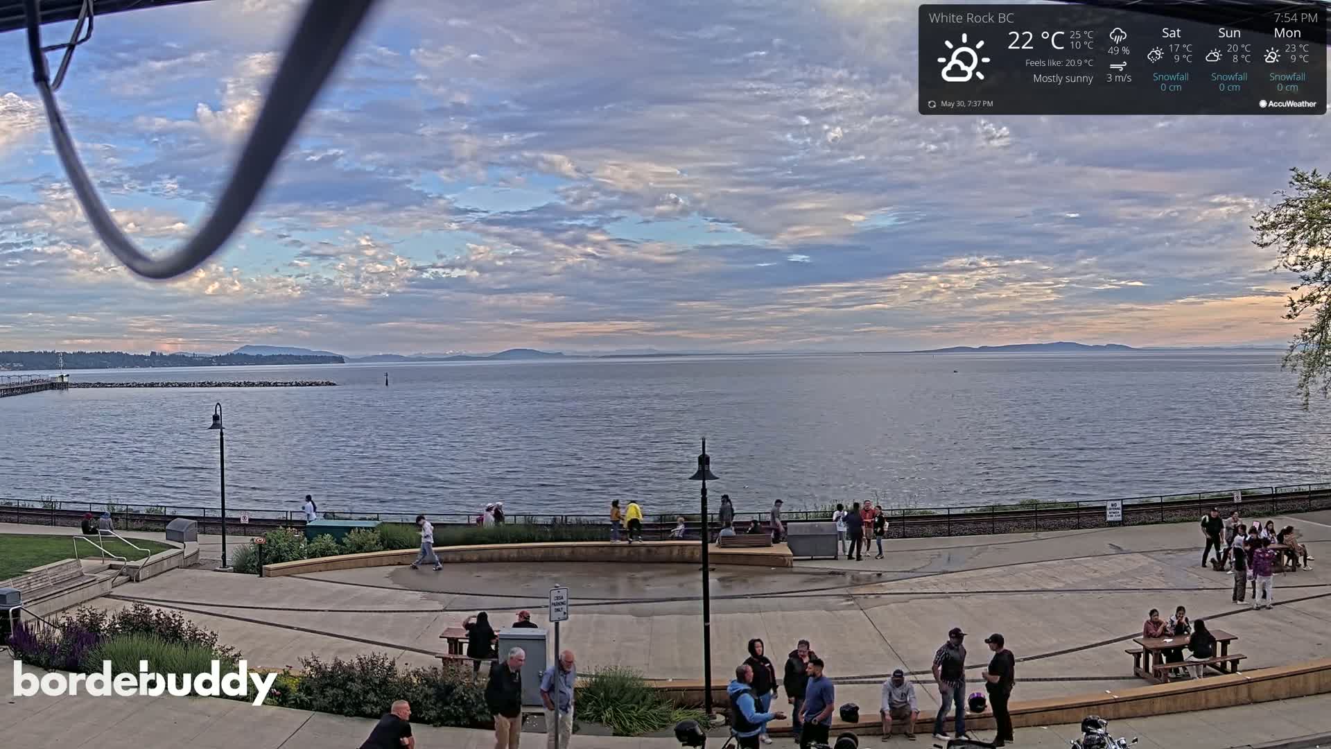 A mostly sunny waterfront scene with calm water, a paved plaza with people gathered in small groups, and a distant mountain range under a partly cloudy sky.