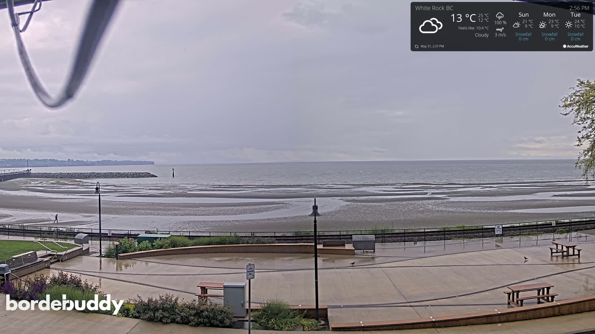 A cloudy, wet day at a seaside plaza shows a wide expanse of tidal flats leading to a calm ocean under an overcast sky.