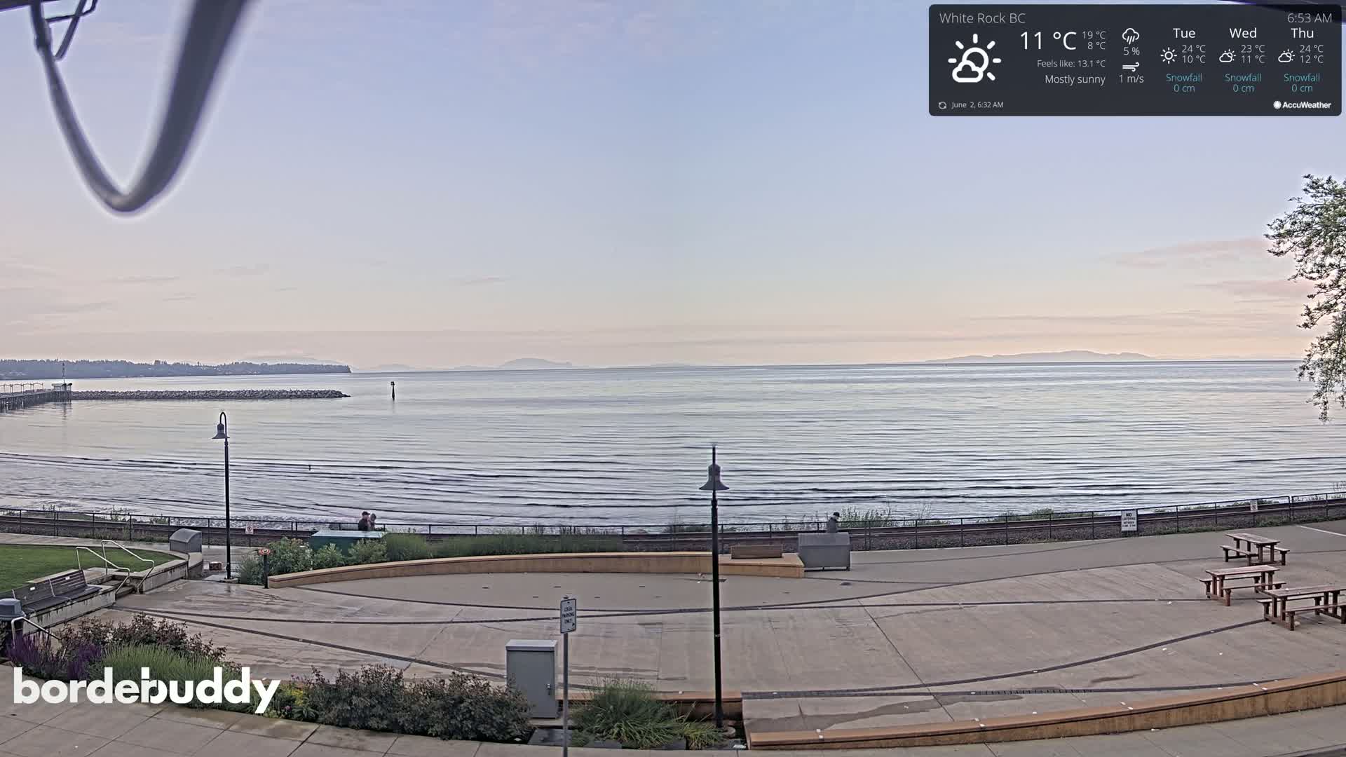 A mostly sunny seaside plaza overlooks a calm ocean with a distant shoreline under a pale sky.