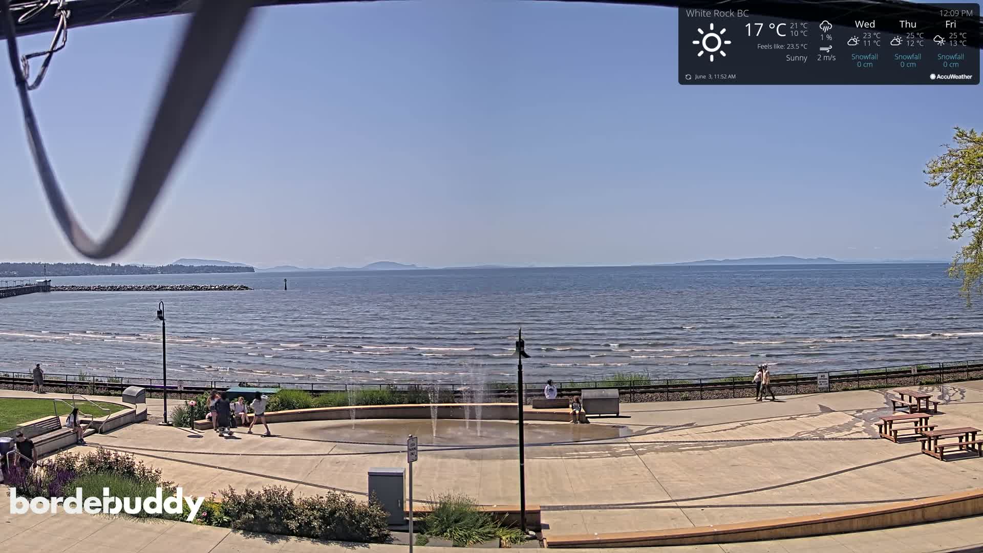 A sunny seaside plaza with a water feature overlooks a calm ocean and distant land under a clear blue sky.