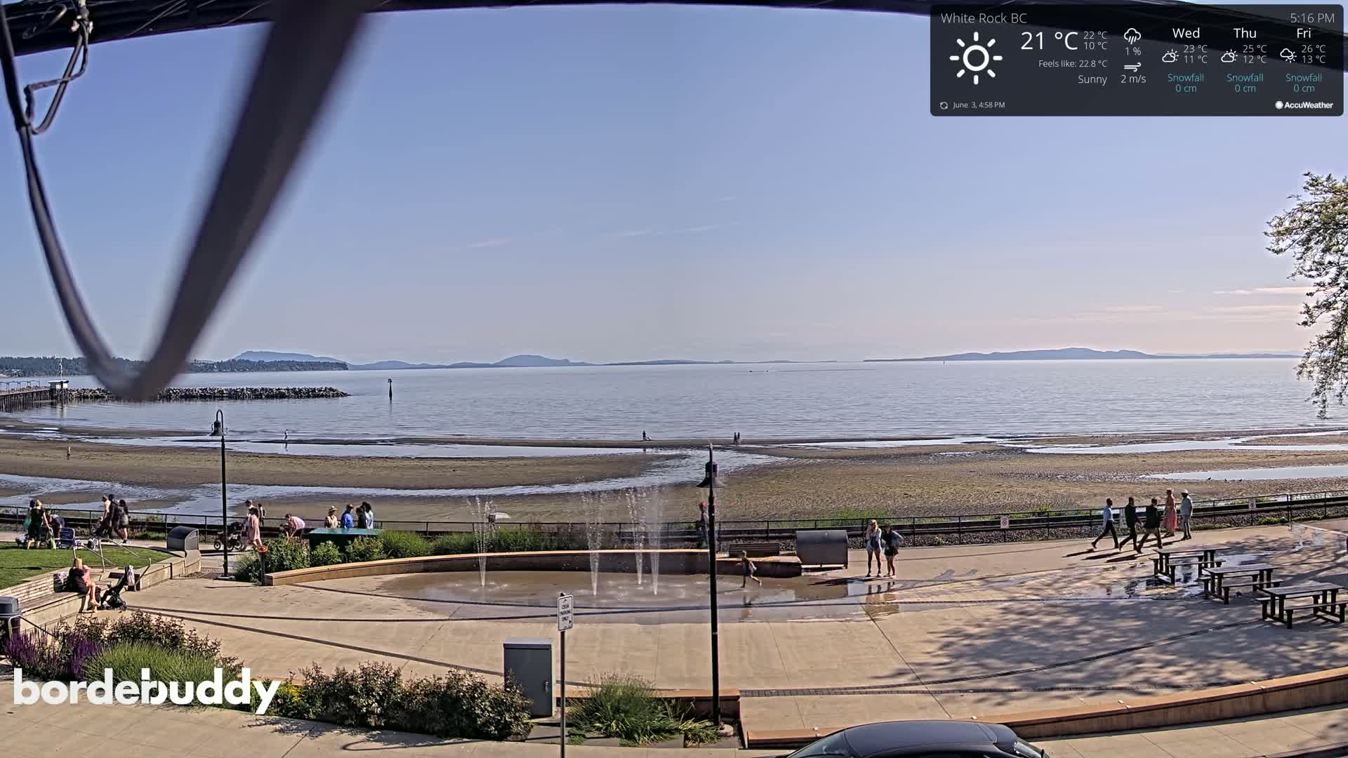 A sunny beach scene shows a calm body of water with distant mountains, a sandy shoreline with people, a water fountain, and picnic tables in a plaza.