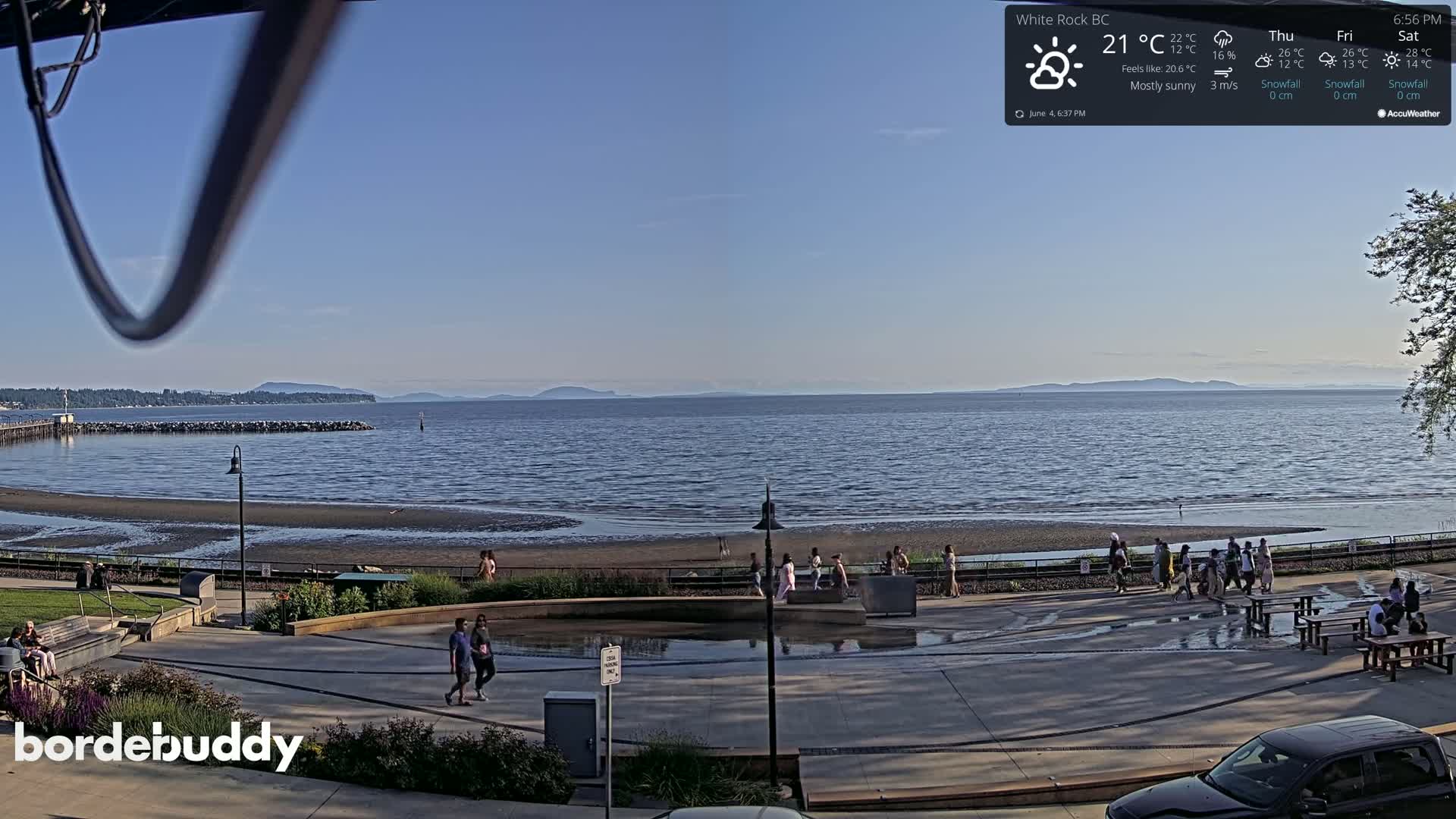 A mostly sunny seaside scene shows a beach with people walking along a paved walkway, overlooking calm water and distant mountains.