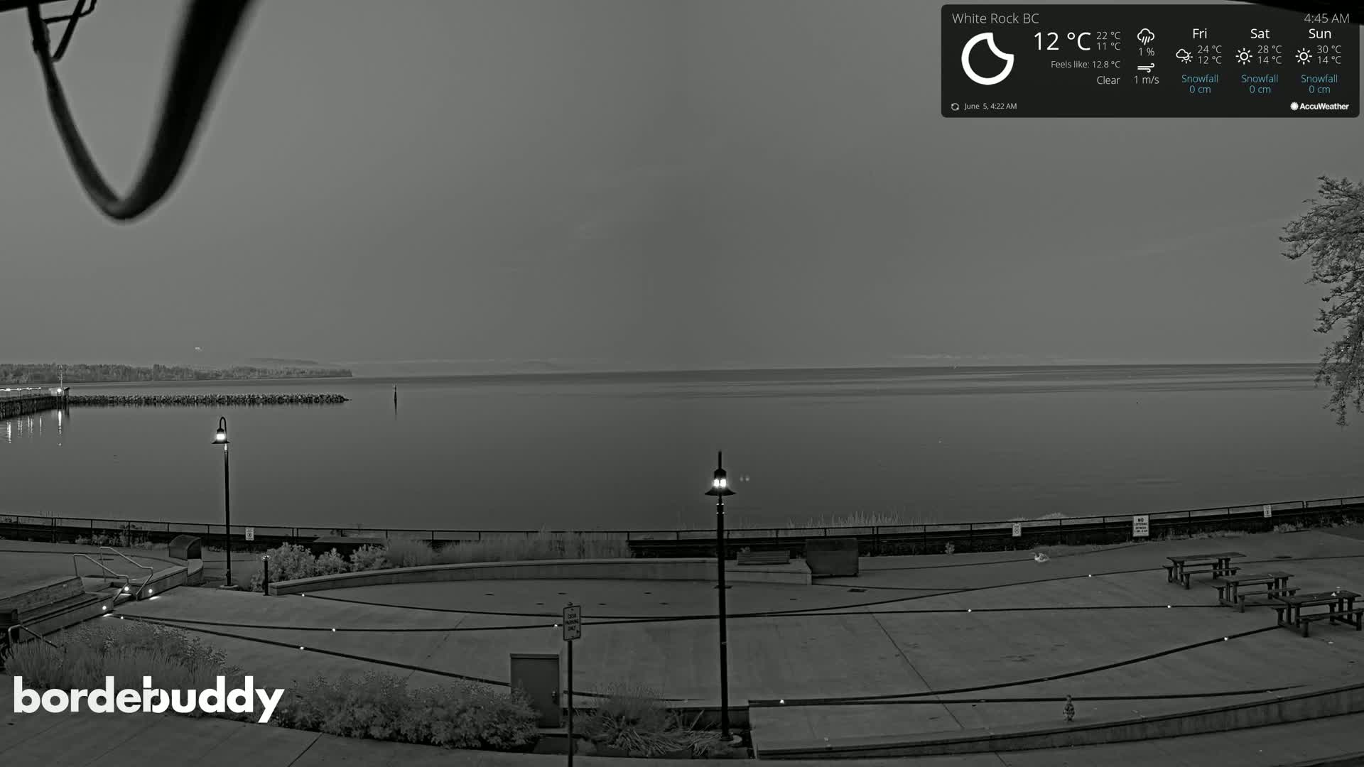 A grayscale image shows a calm body of water at dawn or dusk, with a paved waterfront area containing several picnic tables and lampposts.