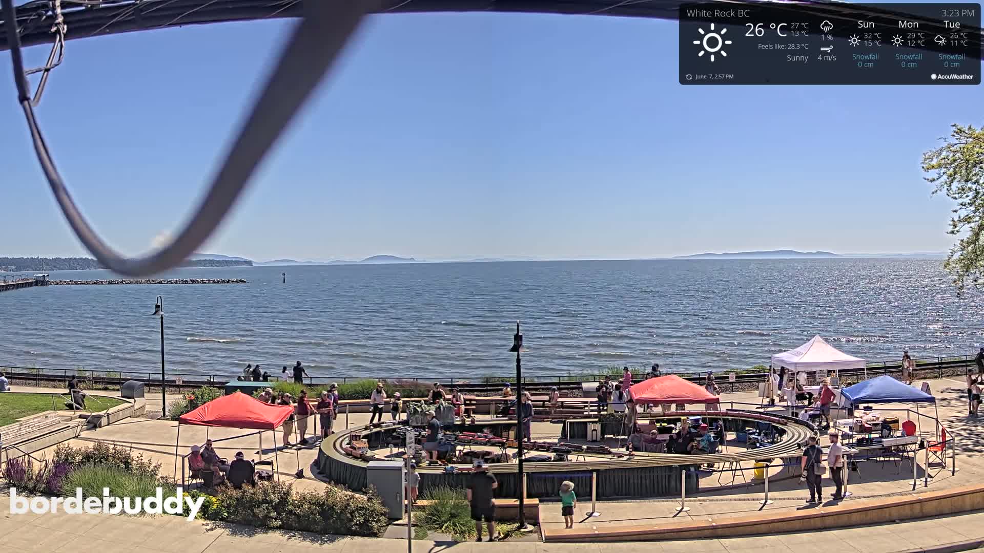 A sunny day overlooks a calm ocean with a shoreline event featuring several tents and a large model train track surrounded by people.