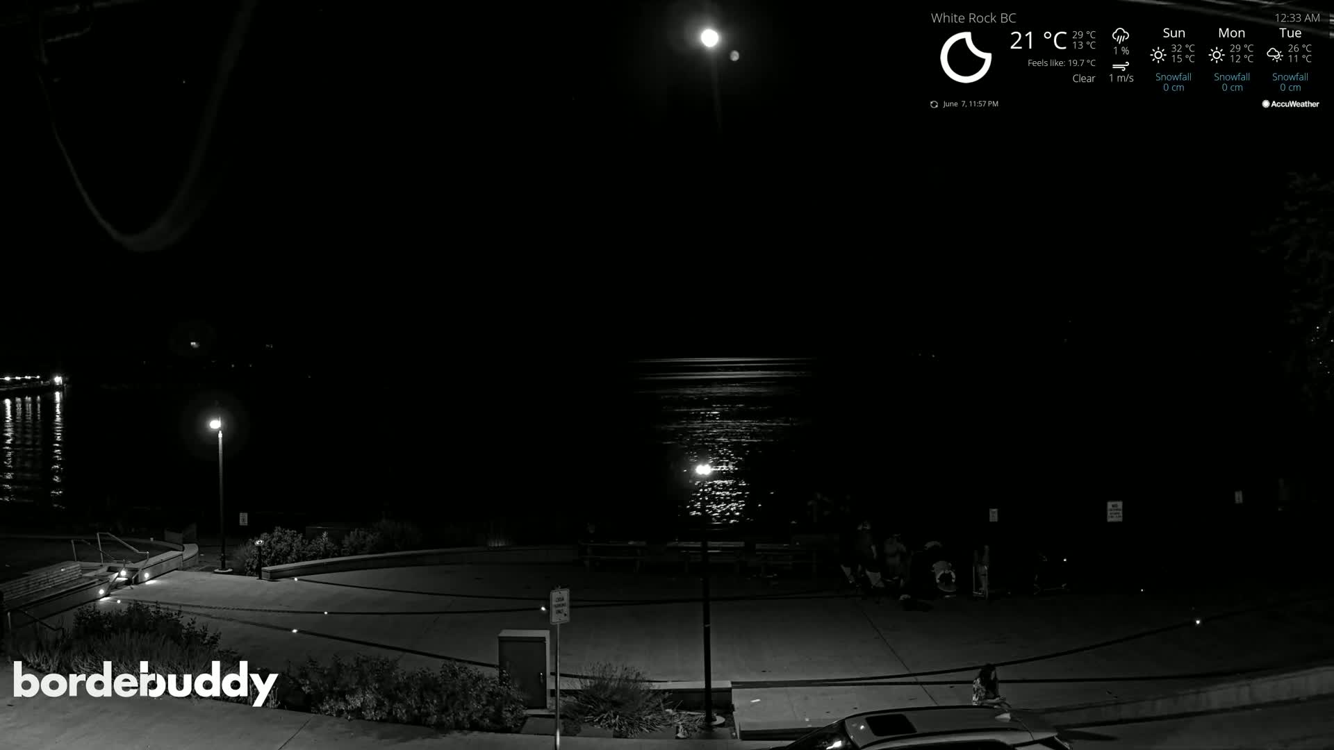 A nighttime view of a waterfront area shows a calm body of water reflecting light under a clear sky, with several people gathered near a paved walkway illuminated by streetlights.