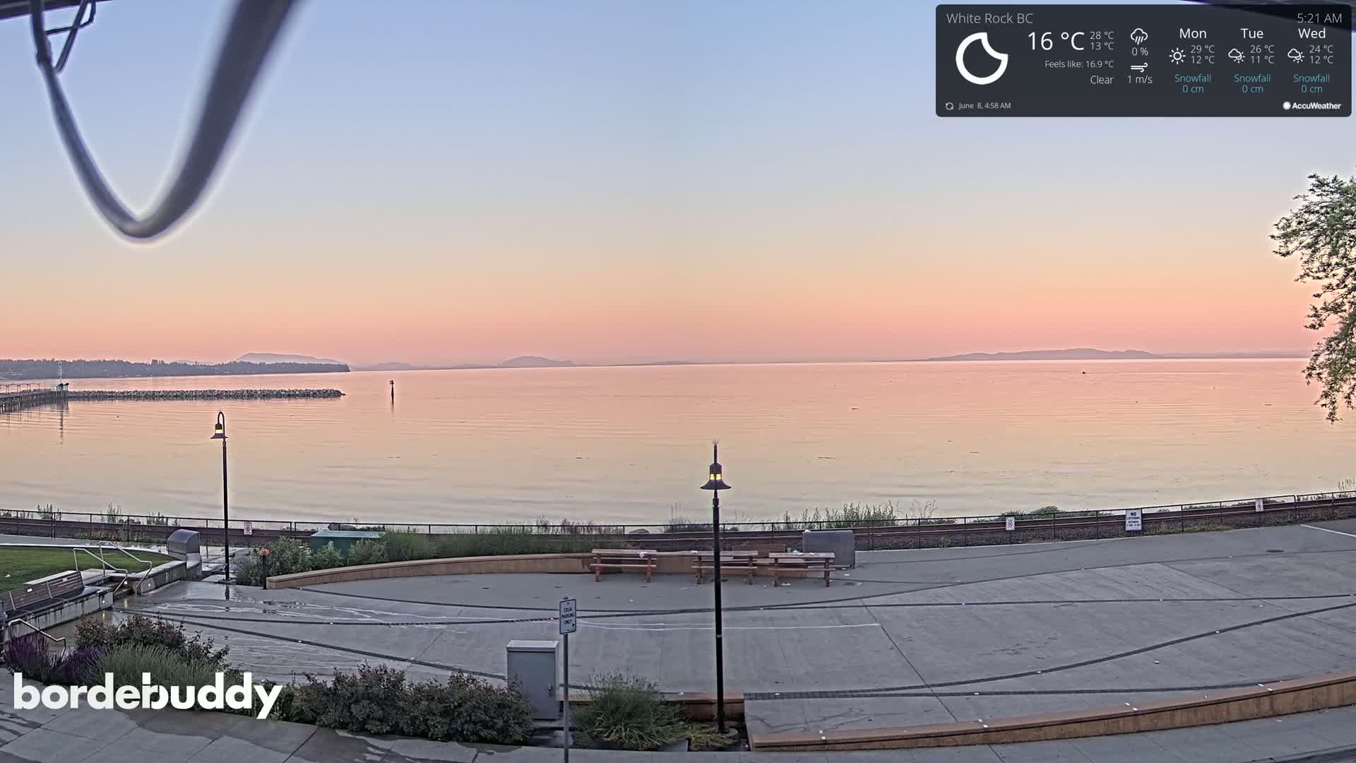 A calm body of water at sunrise, under a clear sky, is viewed from a paved waterfront area with benches and a lamppost.