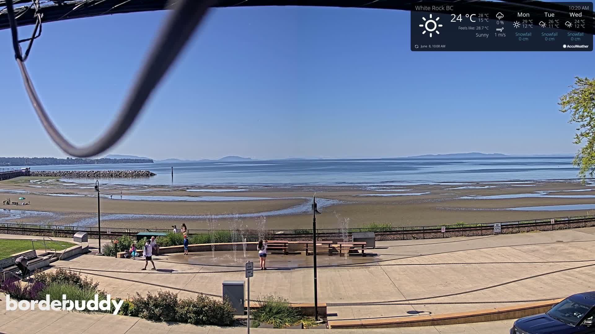 A sunny beach scene shows a wide expanse of calm water receding to reveal a sandy shoreline with several people enjoying a water feature near picnic tables and a paved walkway.