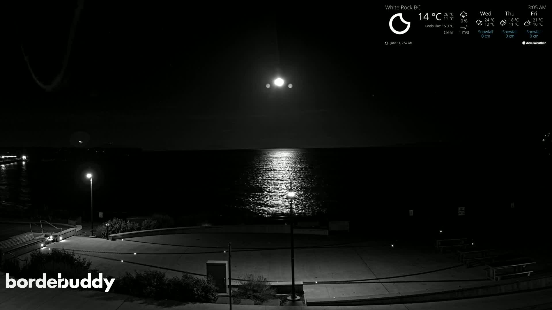 A bright moon shines over a calm body of water at night, illuminating a dark, empty plaza with a few lights.