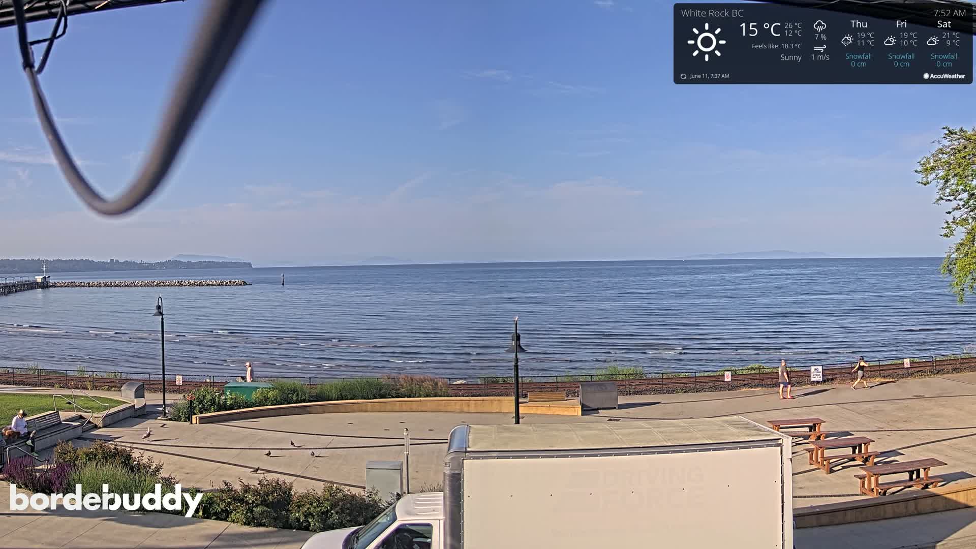 A sunny seaside view shows calm ocean waves lapping a shoreline with a paved walkway, picnic tables, and a few people, all under a clear blue sky.
