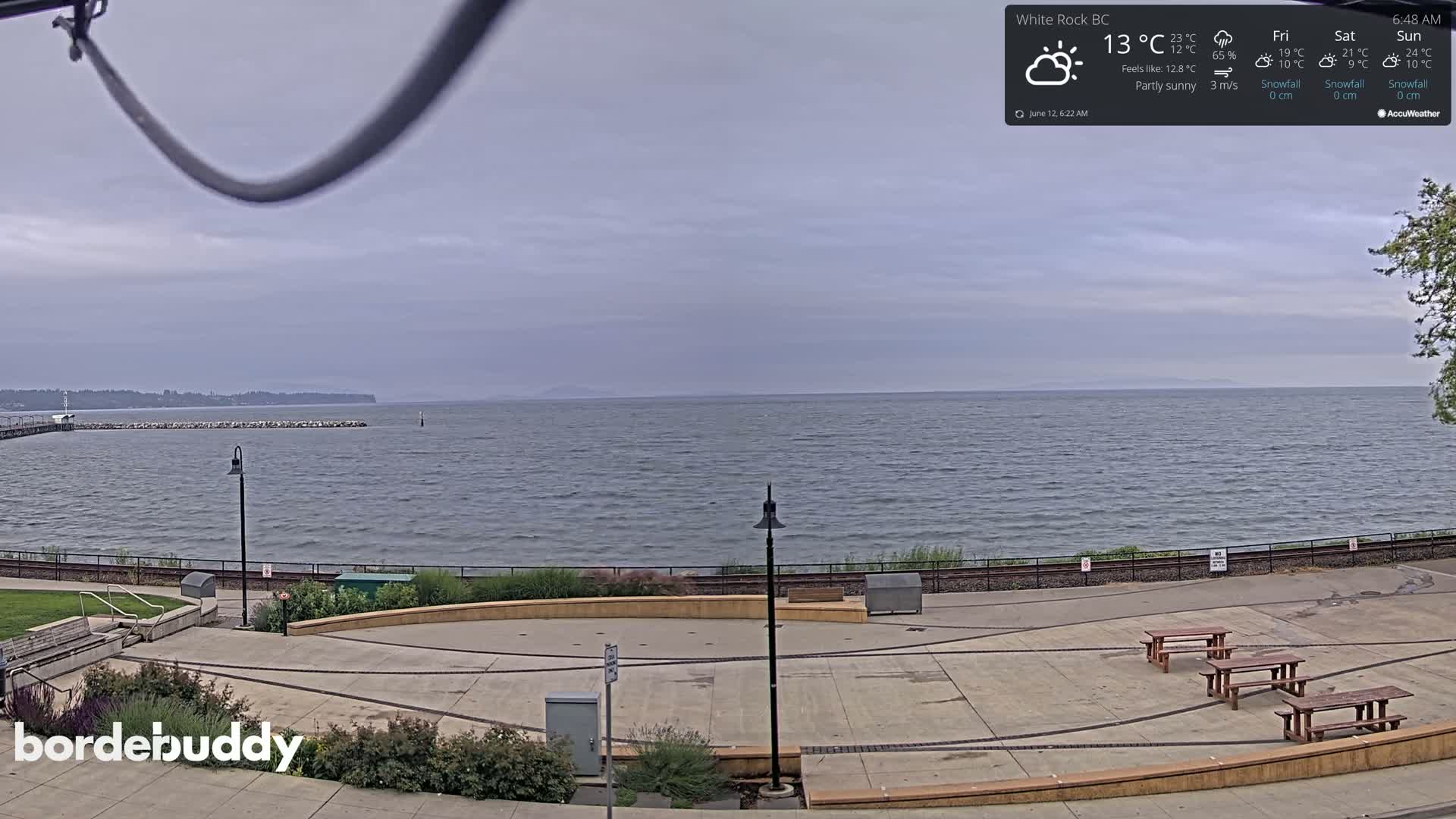A partly sunny waterfront scene shows a paved plaza with benches overlooking a calm body of water and a distant shoreline.