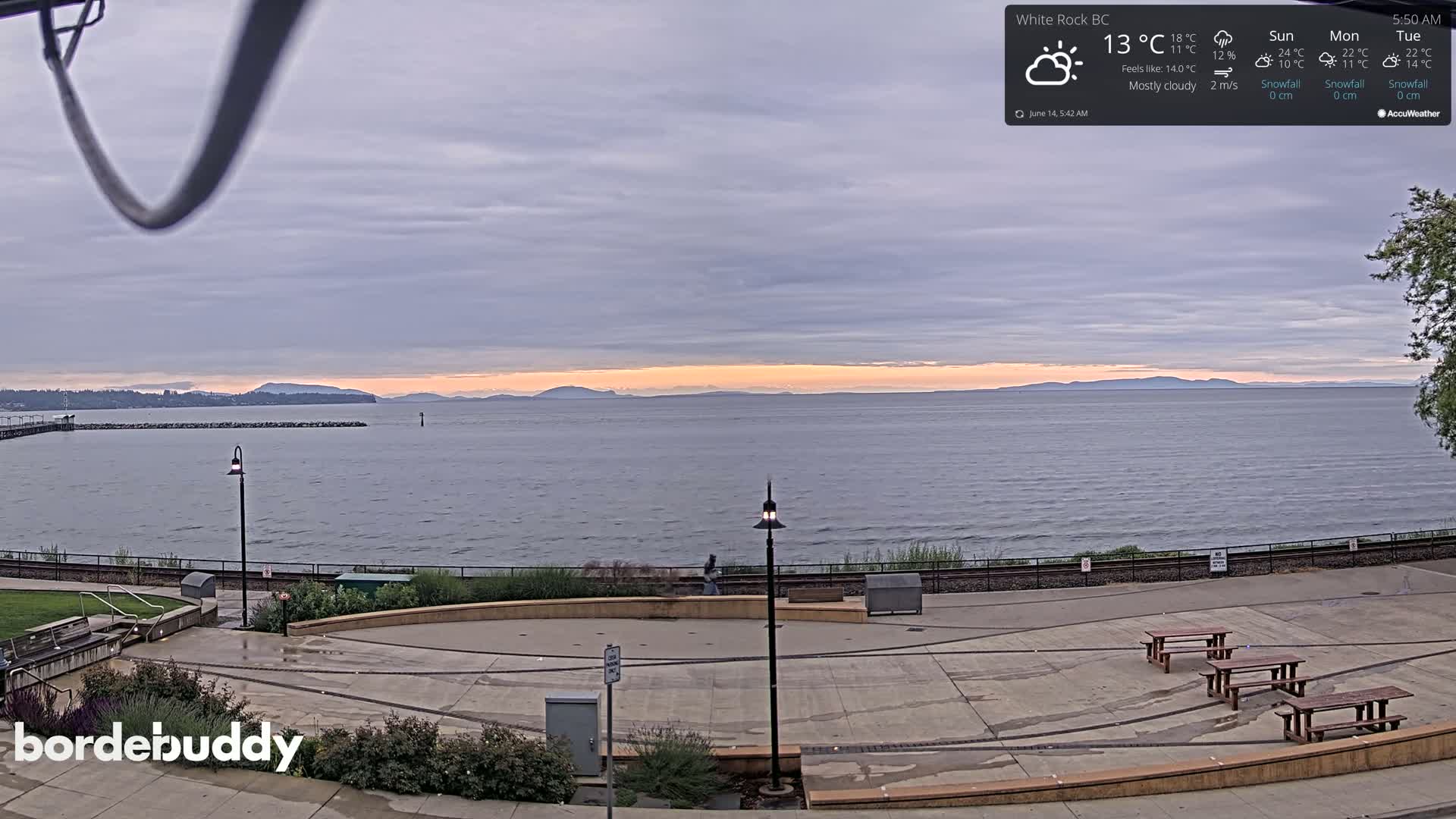 A mostly cloudy sunrise over a calm body of water, viewed from a paved waterfront area with benches and lampposts.