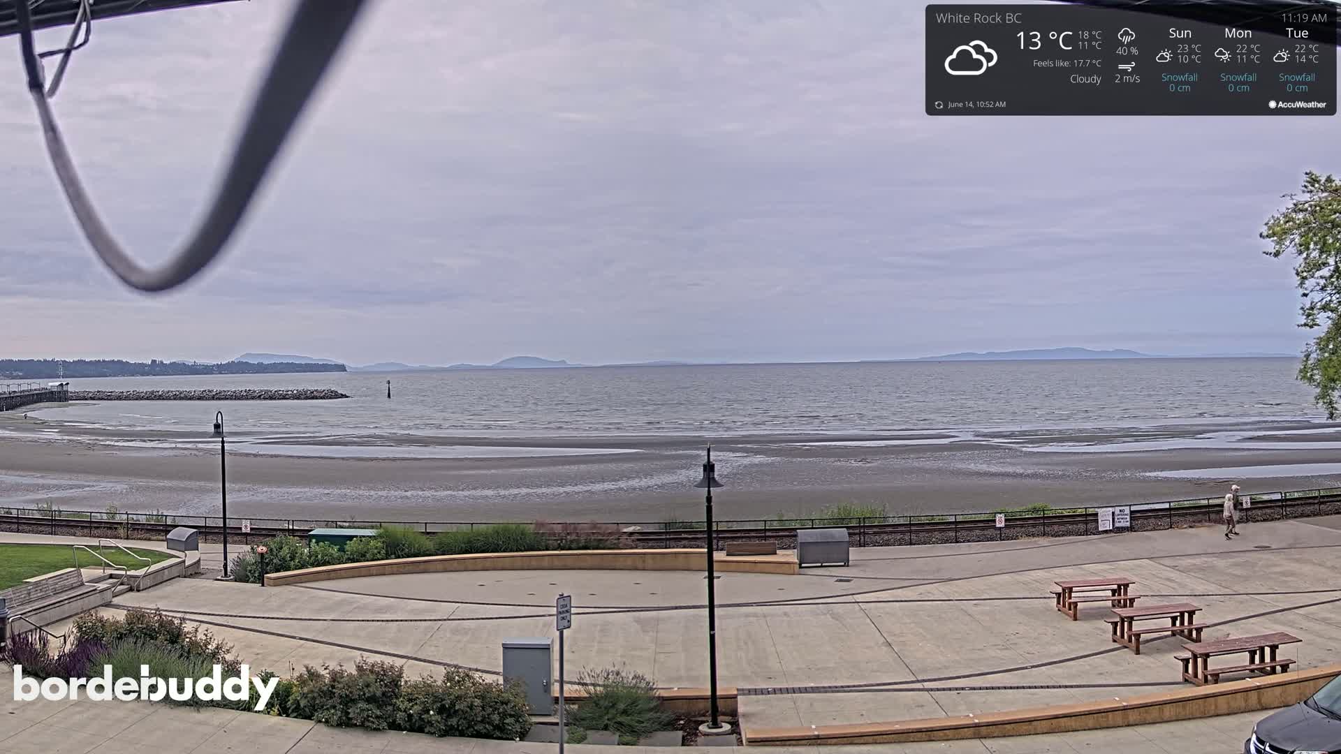 A cloudy day overlooking a wide, sandy beach and calm ocean with distant mountains, and a paved plaza with picnic tables in the foreground.