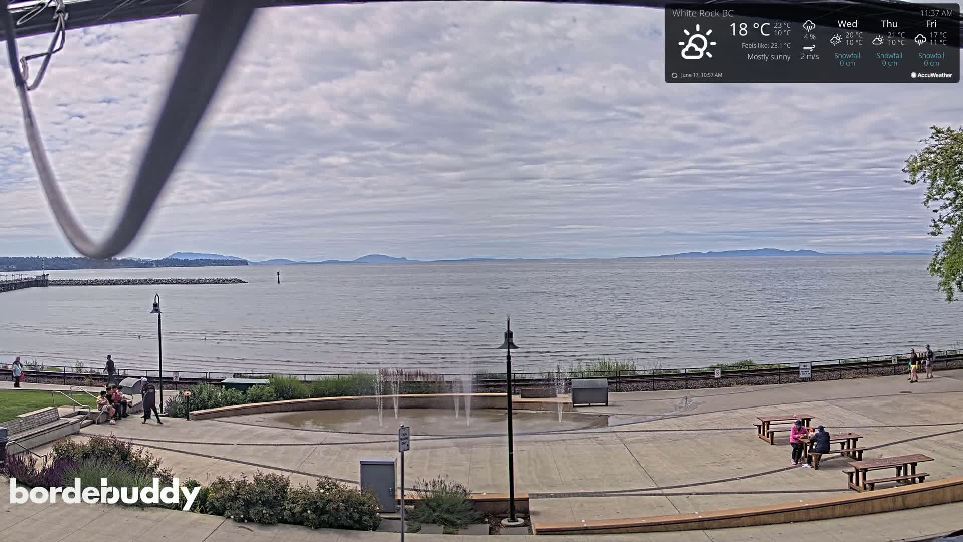 A mostly sunny waterfront scene shows a calm body of water with distant mountains under a partly cloudy sky, featuring a paved plaza with small fountains and a few people scattered around.