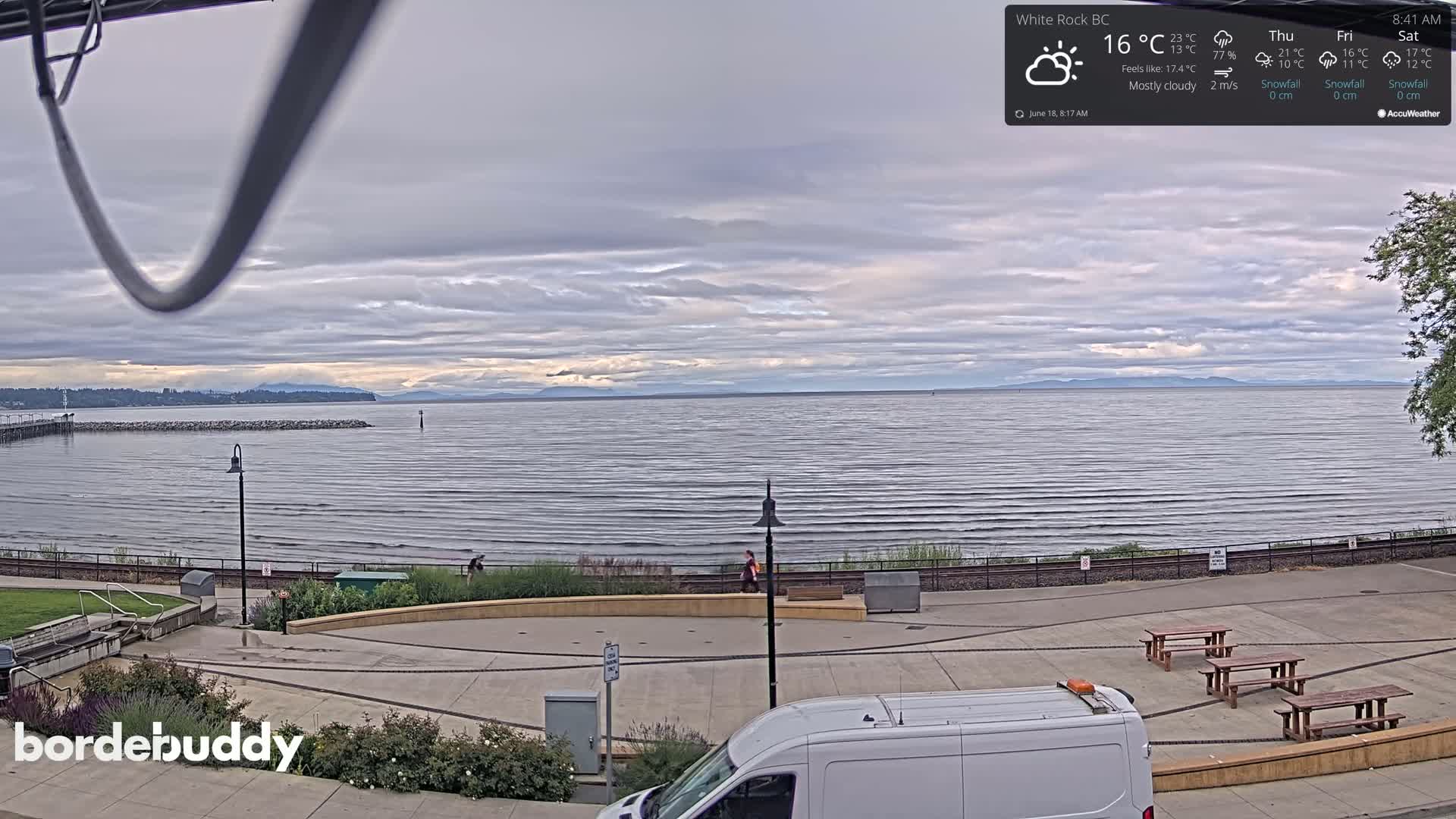 A mostly cloudy day shows a calm body of water with a shoreline featuring paved walkways, benches, and a white van; mountains are visible in the distance.
