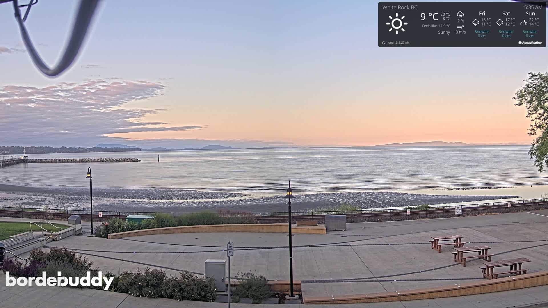 A calm, sunny seaside scene at dawn shows a paved waterfront area with picnic tables overlooking a wide expanse of calm water and distant mountains under a pale pink sky.