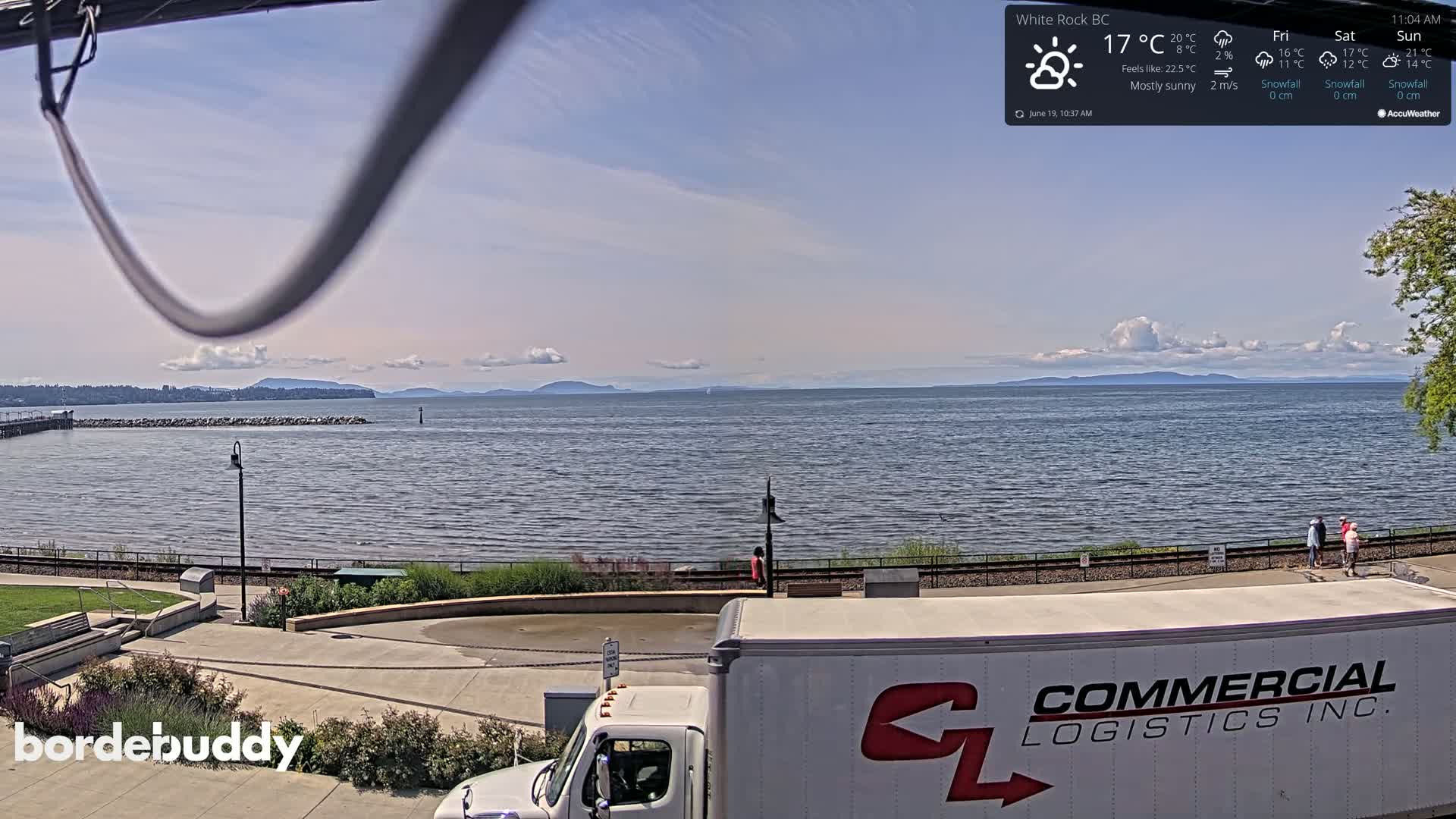 A mostly sunny waterfront scene shows a calm body of water with distant mountains under a partly cloudy sky, a paved walkway, and a large white delivery truck.