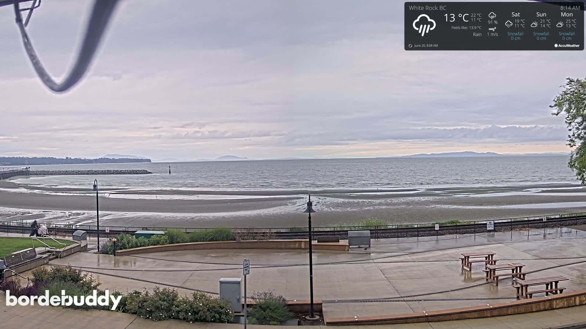 A wet, overcast beach scene shows a wide expanse of sand and calm water under a gray sky, with a paved area containing picnic tables in the foreground.