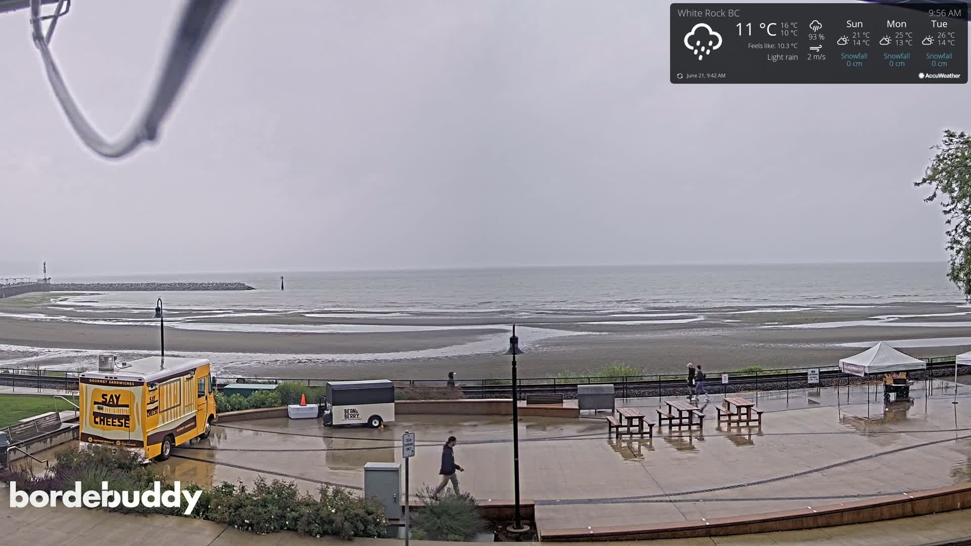 A wet, grey day at a beachside plaza with food trucks, picnic tables, and a few people walking; light rain is falling over a calm sea.