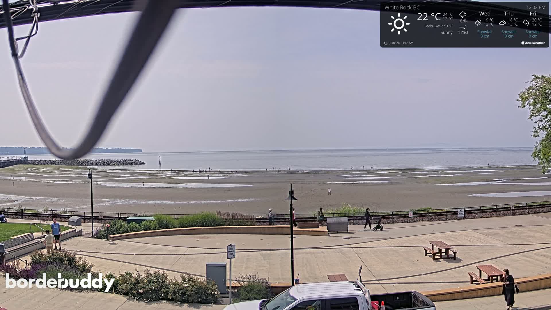 A sunny beach with a calm ocean stretches out before a paved promenade with people strolling along it.