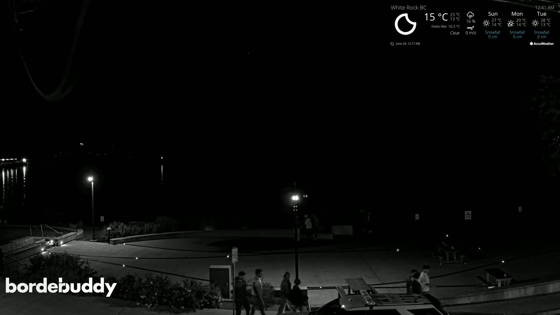 At night, under a clear sky, several people walk across a paved area near a body of water, which is dimly lit by several lamps.