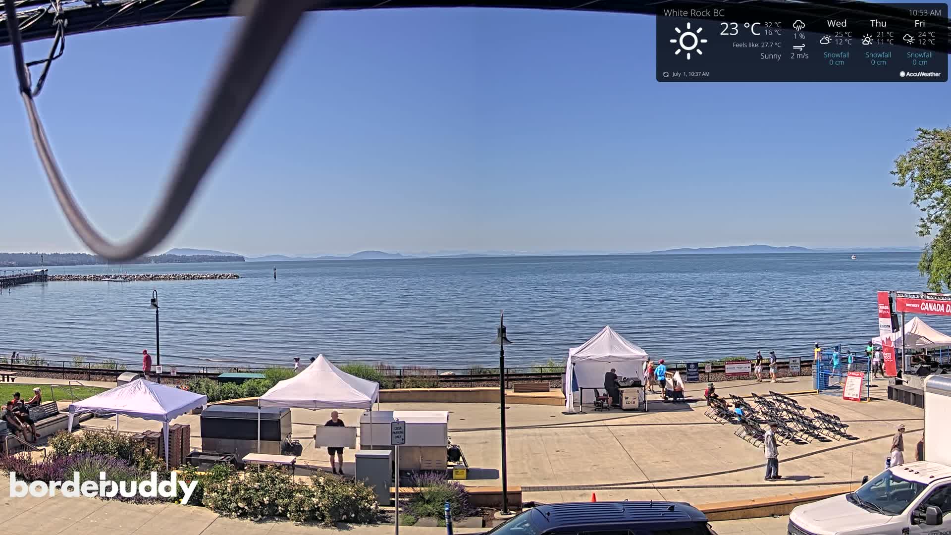 A sunny waterfront scene features several white tents, folding chairs, and parked vehicles in a plaza overlooking a calm body of water with distant mountains under a clear sky.