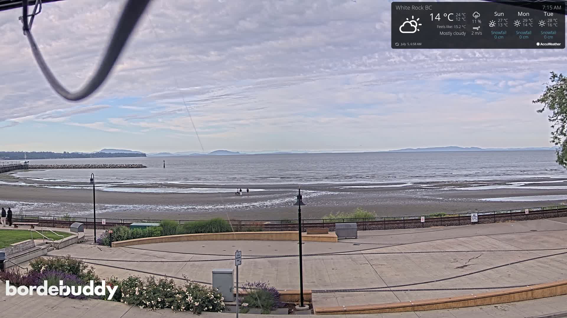 A mostly cloudy view of a beach with a calm ocean and distant mountains, overlooking a paved plaza.