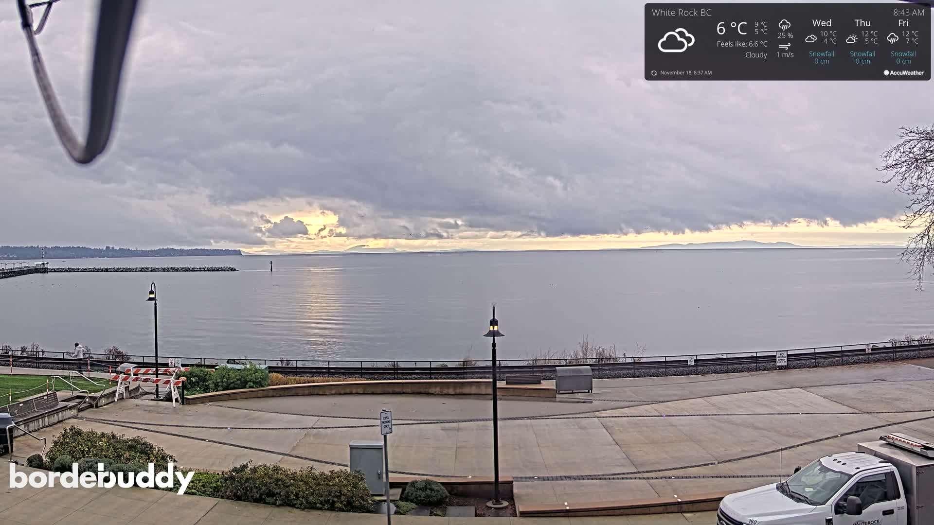A mostly cloudy night scene shows a dark body of water reflecting lights, and a paved area with some people and a vehicle, lit by several lamps.