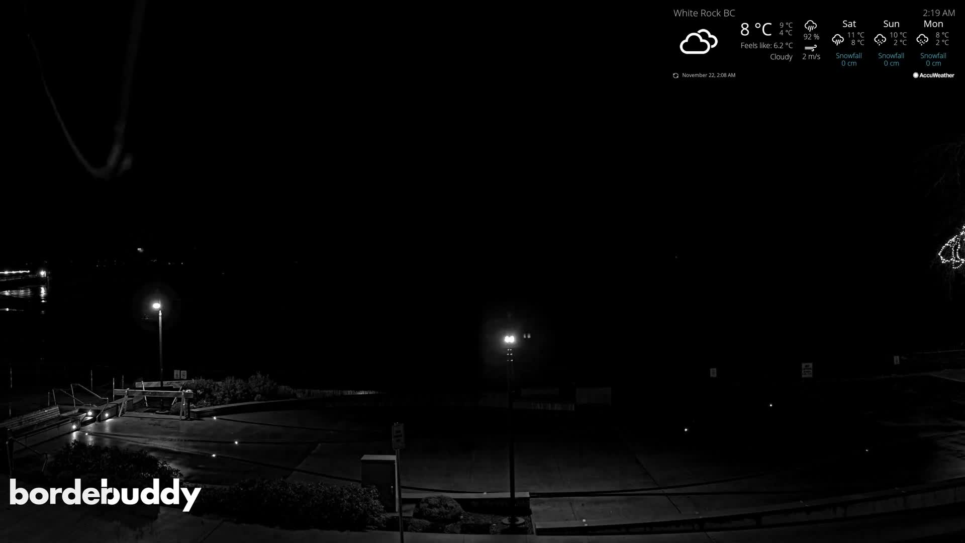 A dark, calm night scene shows a brightly lit streetlamp on a waterfront promenade with benches, next to still water reflecting distant lights.