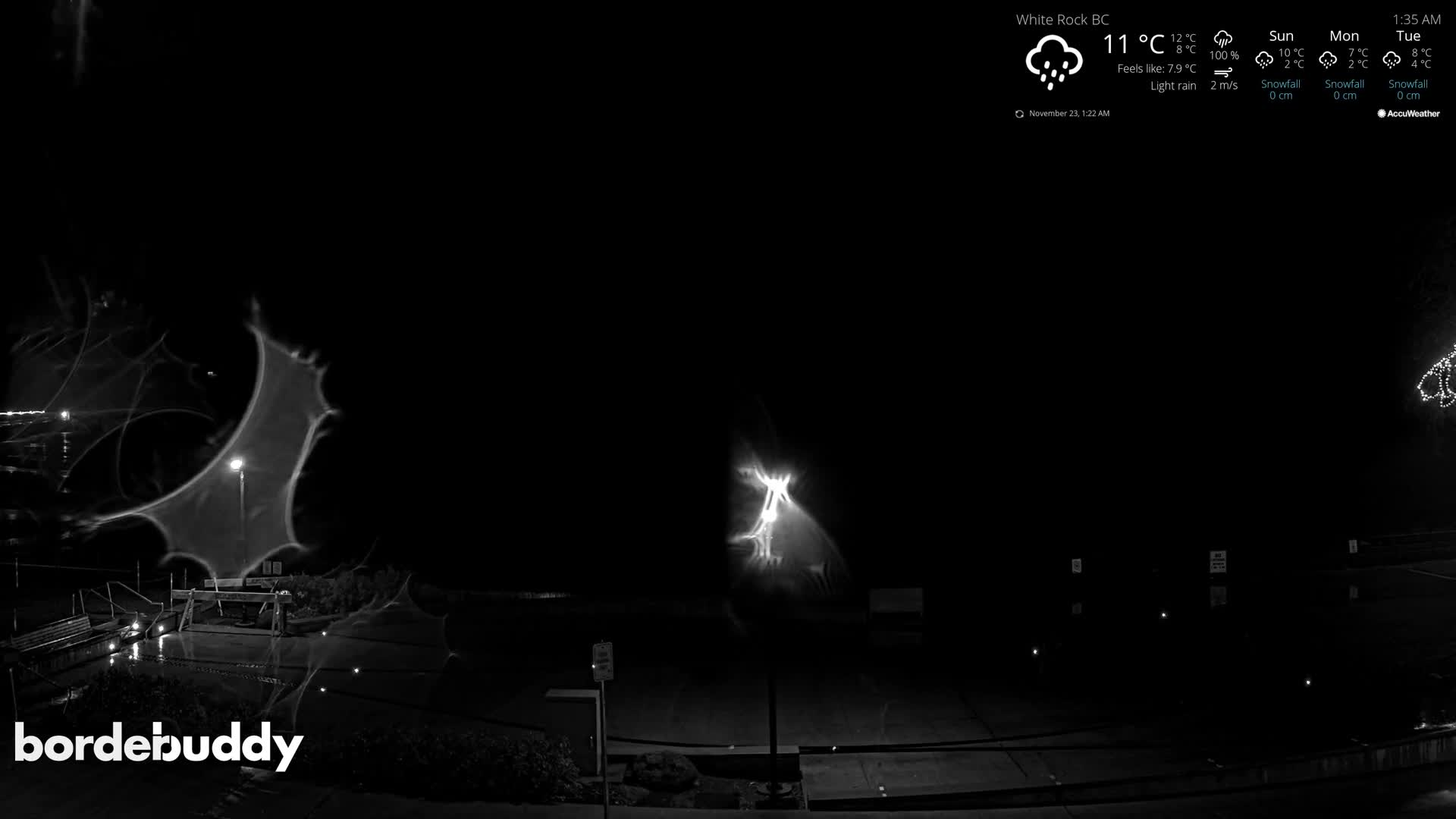 The image shows a dark, rainy outdoor scene at night, with bright streetlights reflecting on wet ground and prominent abstract light patterns, possibly from debris or moisture on the camera lens.