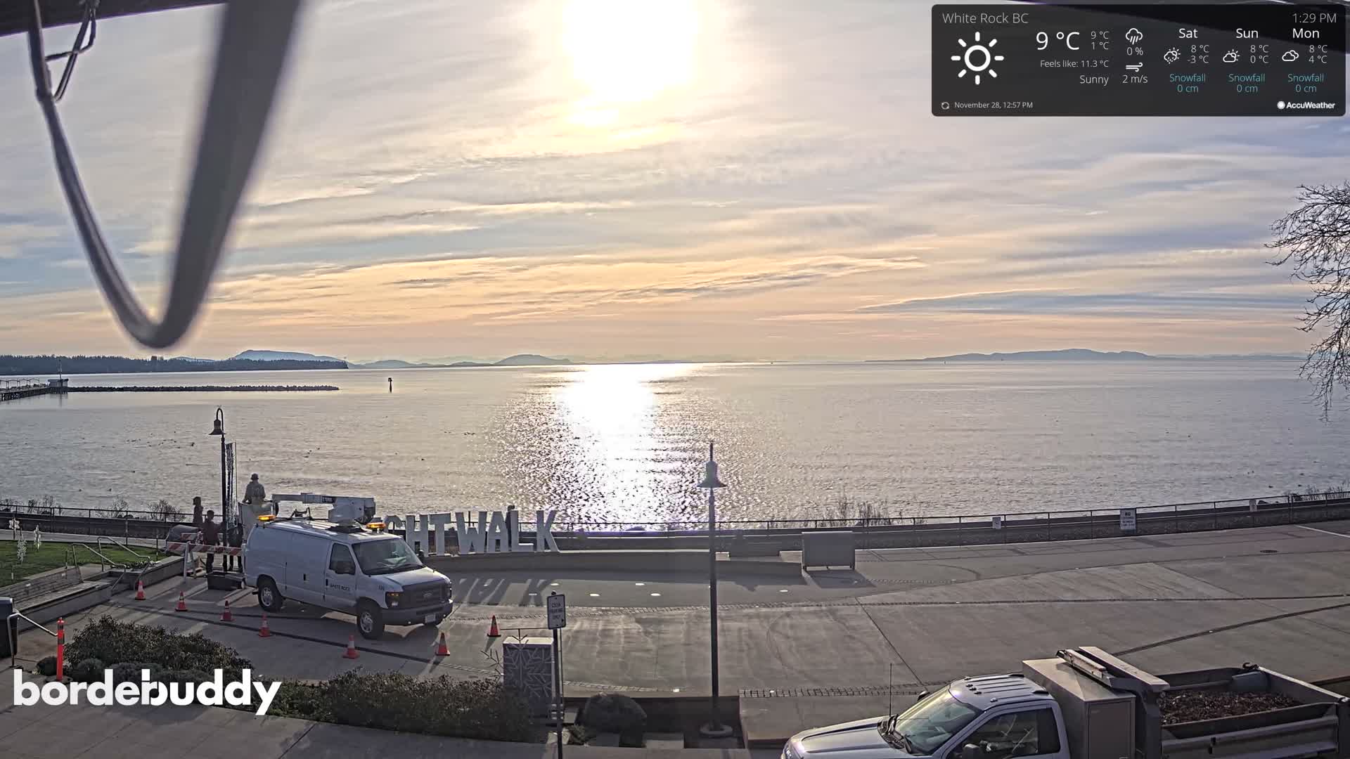 On a bright, sunny day with scattered clouds, a utility van with workers on a cherry picker is parked near large letters along a waterfront promenade, overlooking glistening water with distant land and a pier under a partly cloudy sky.