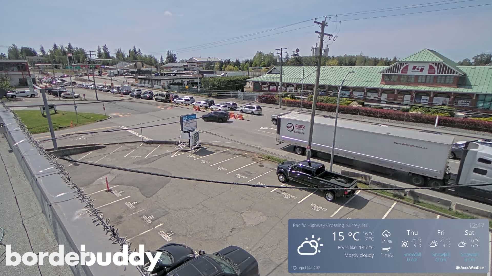 A mostly cloudy day shows a parking lot and roadway with several cars and trucks, some of which are stopped in a line.