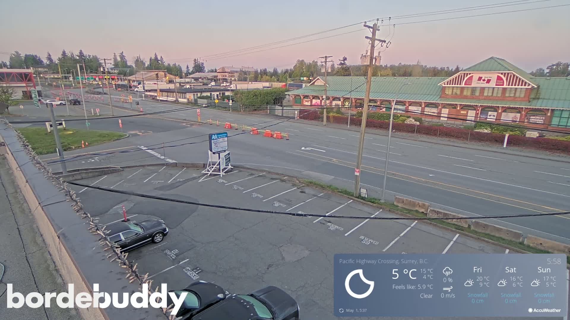 A mostly empty parking lot is situated next to a multi-lane road with a large building across the street, under a clear sky at dawn.