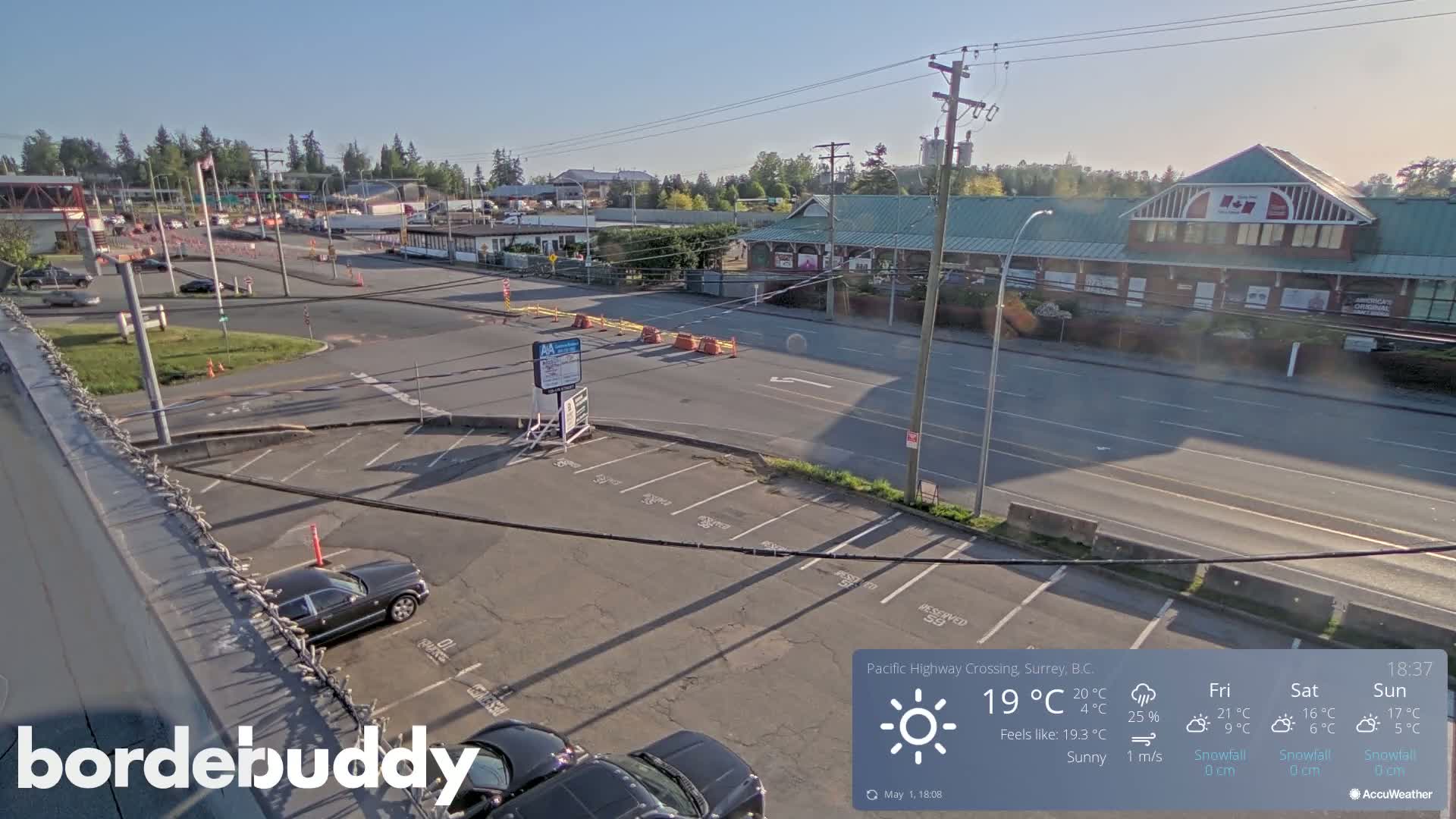 A sunny overhead view of a roadway intersection with a parking lot, buildings, and sparse traffic.