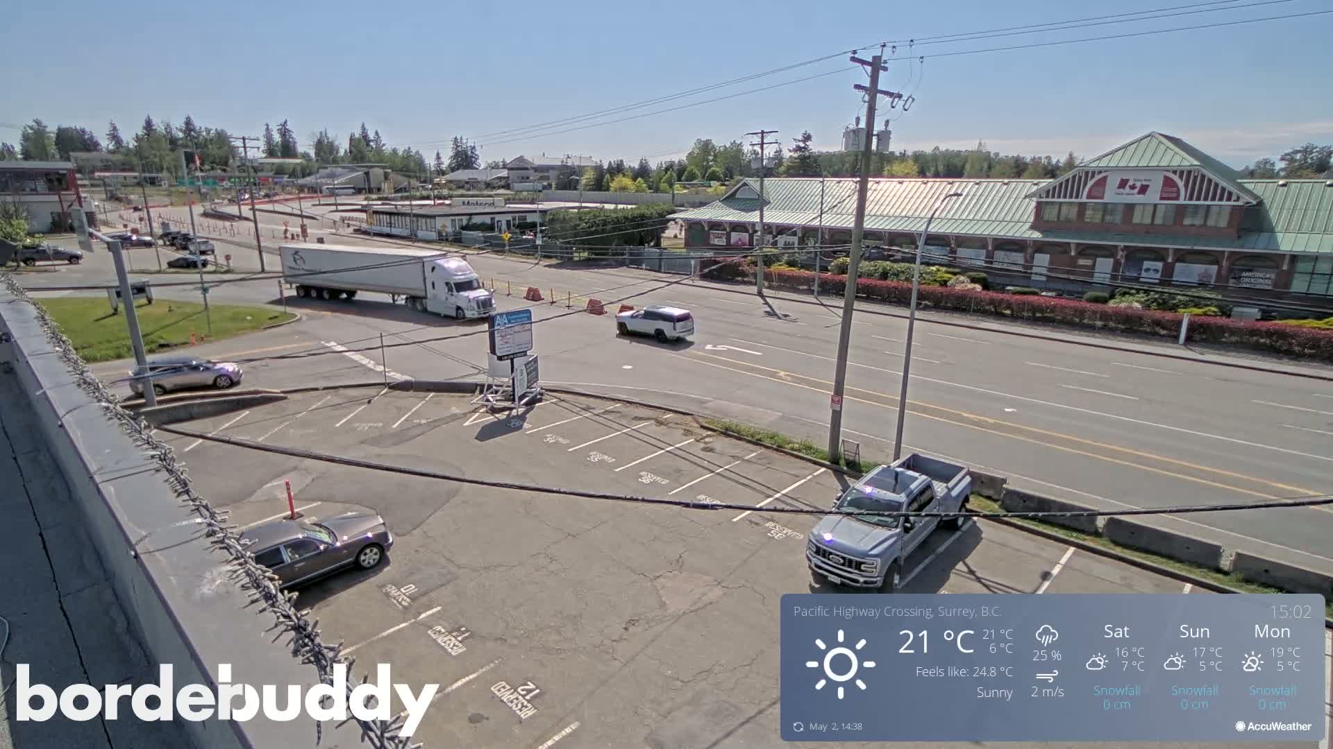 A sunny overhead view of a parking lot and roadway, with several vehicles, including a semi-truck and pickup truck, and a large building in the background.