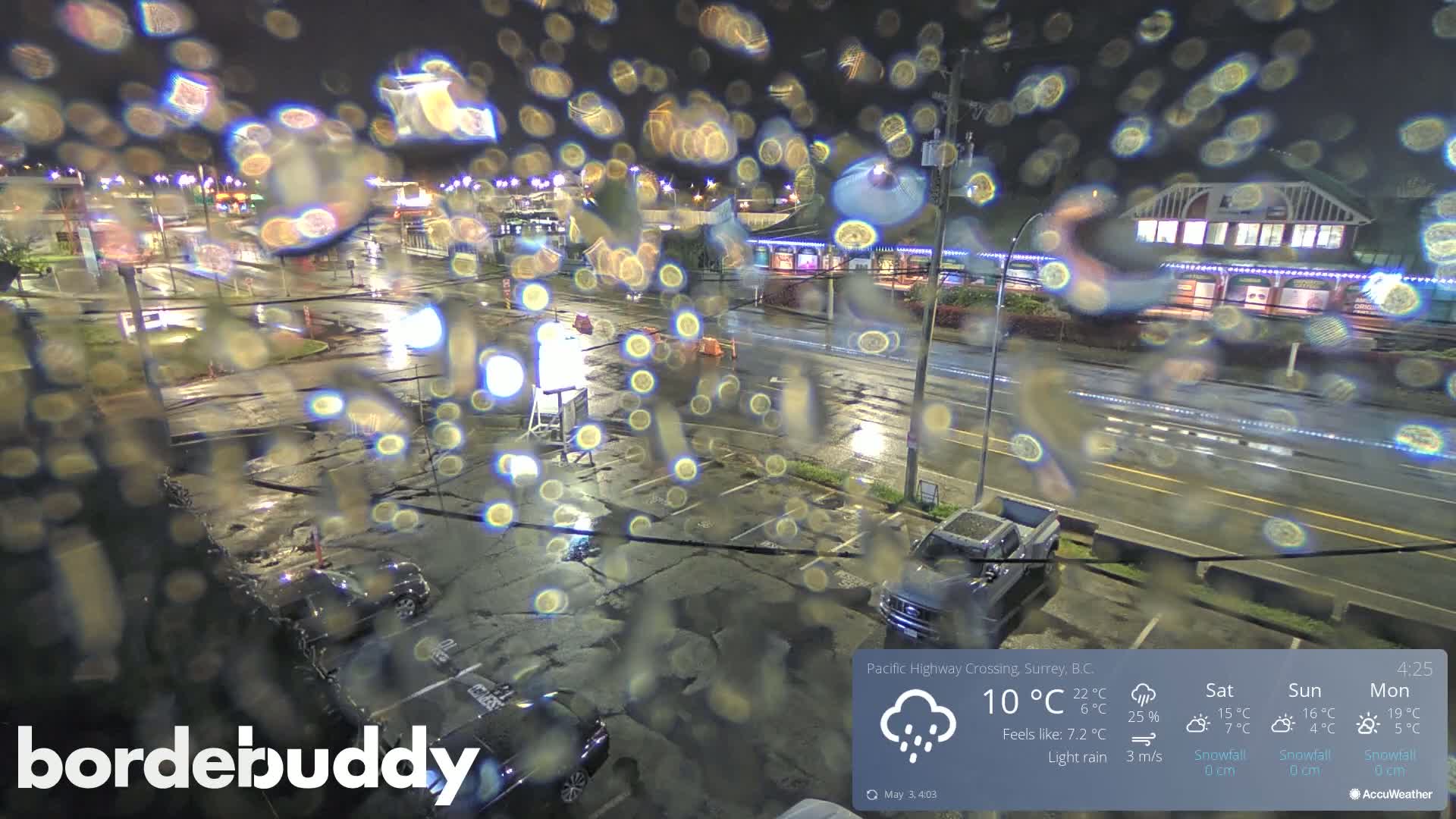 A rain-streaked nighttime view of a wet parking lot and road, with cars and buildings visible in the background under light rain.
