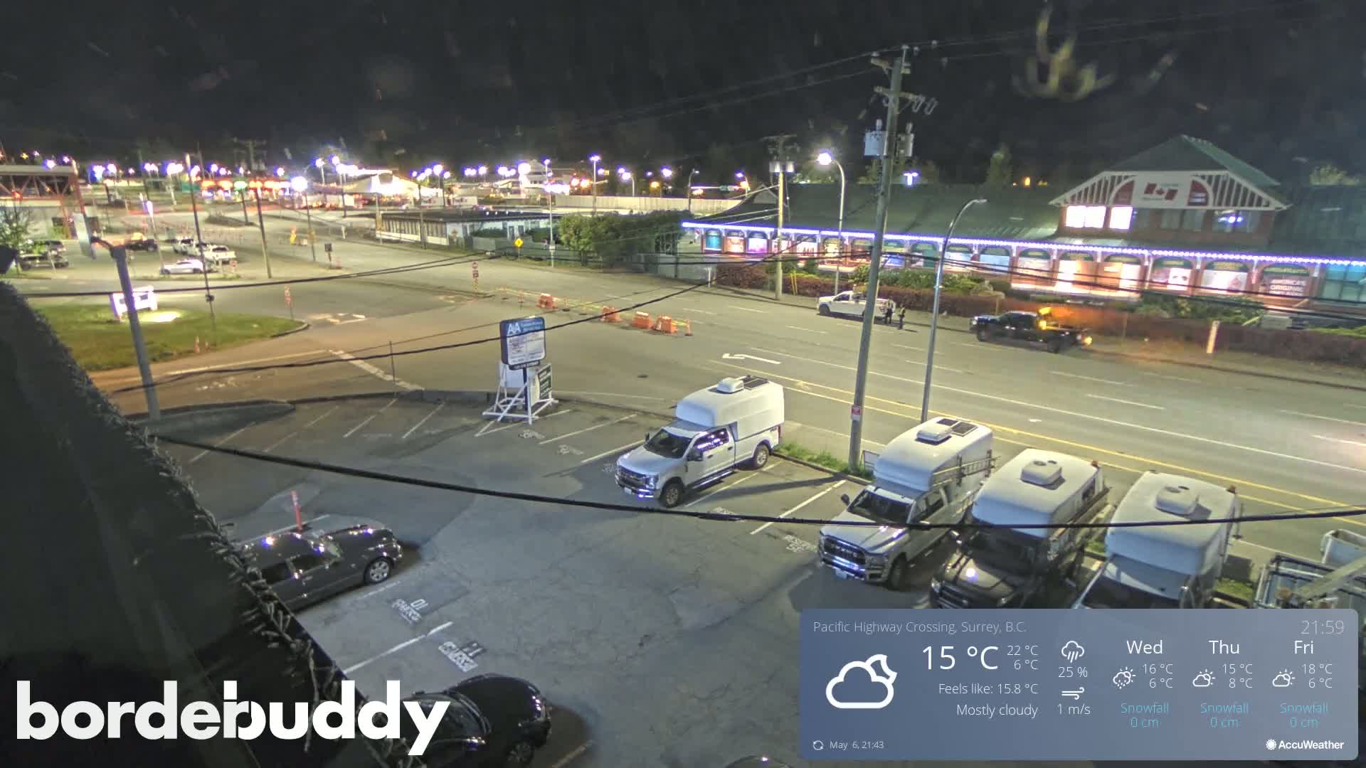 A mostly cloudy night scene shows a parking lot with several vehicles, including trucks and RVs, next to a road with cars and a building with lights.