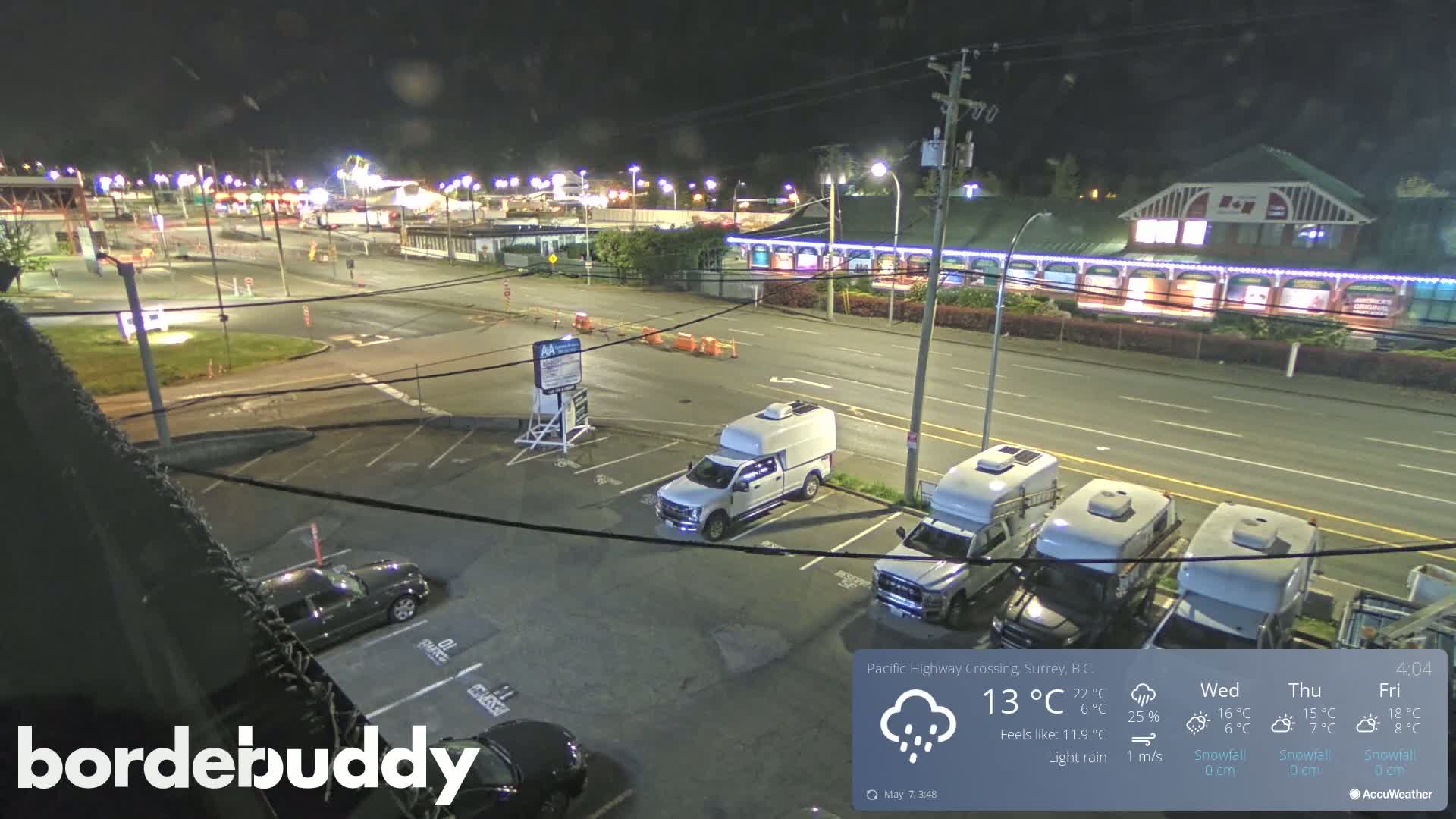 A nighttime, lightly raining scene shows a parking lot with several vehicles, including a pickup truck and RVs, next to a roadway with illuminated buildings in the background.