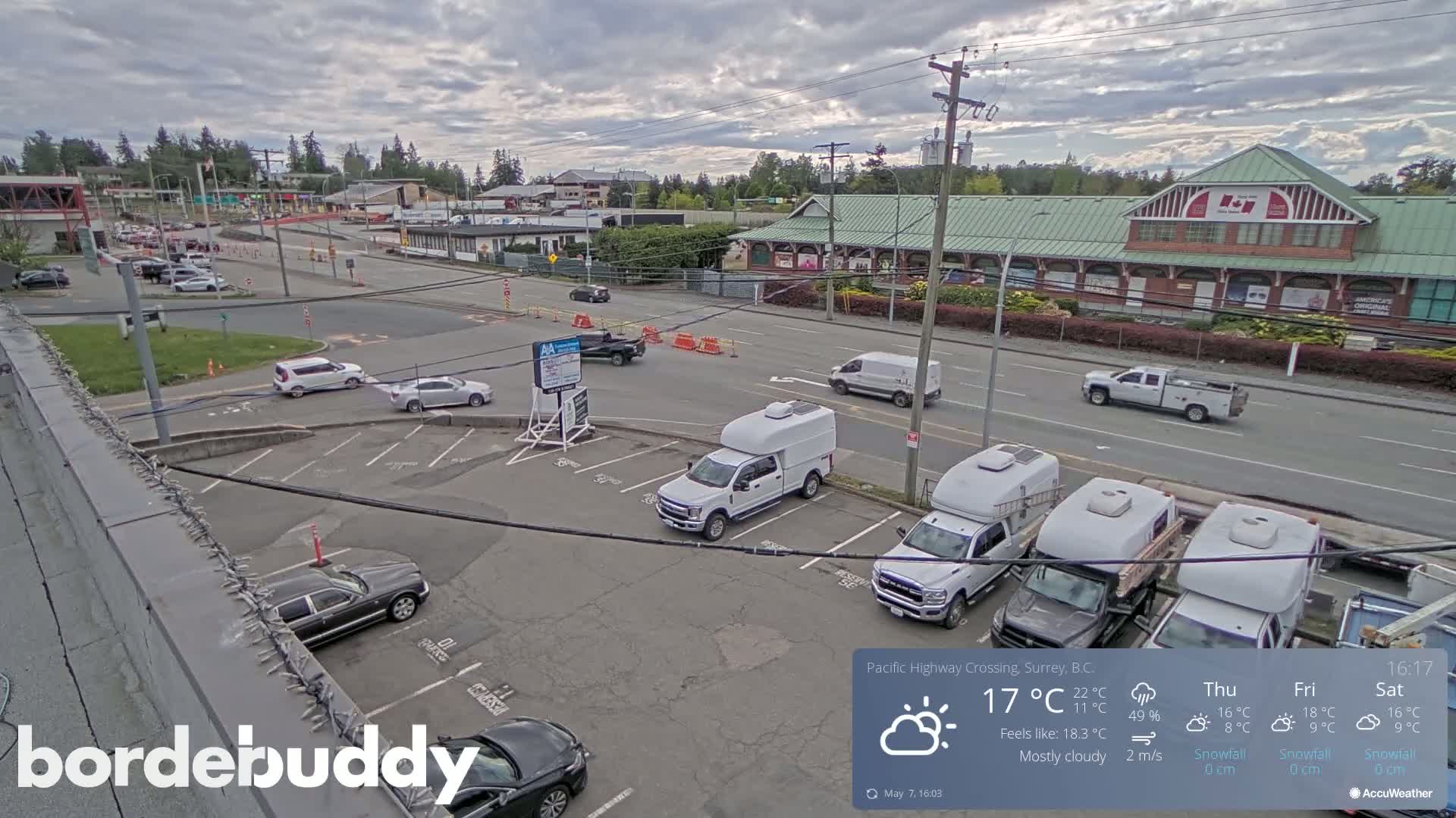 A mostly cloudy day shows a parking lot with several cars and trucks, a roadway with some vehicles, and a building in the background.