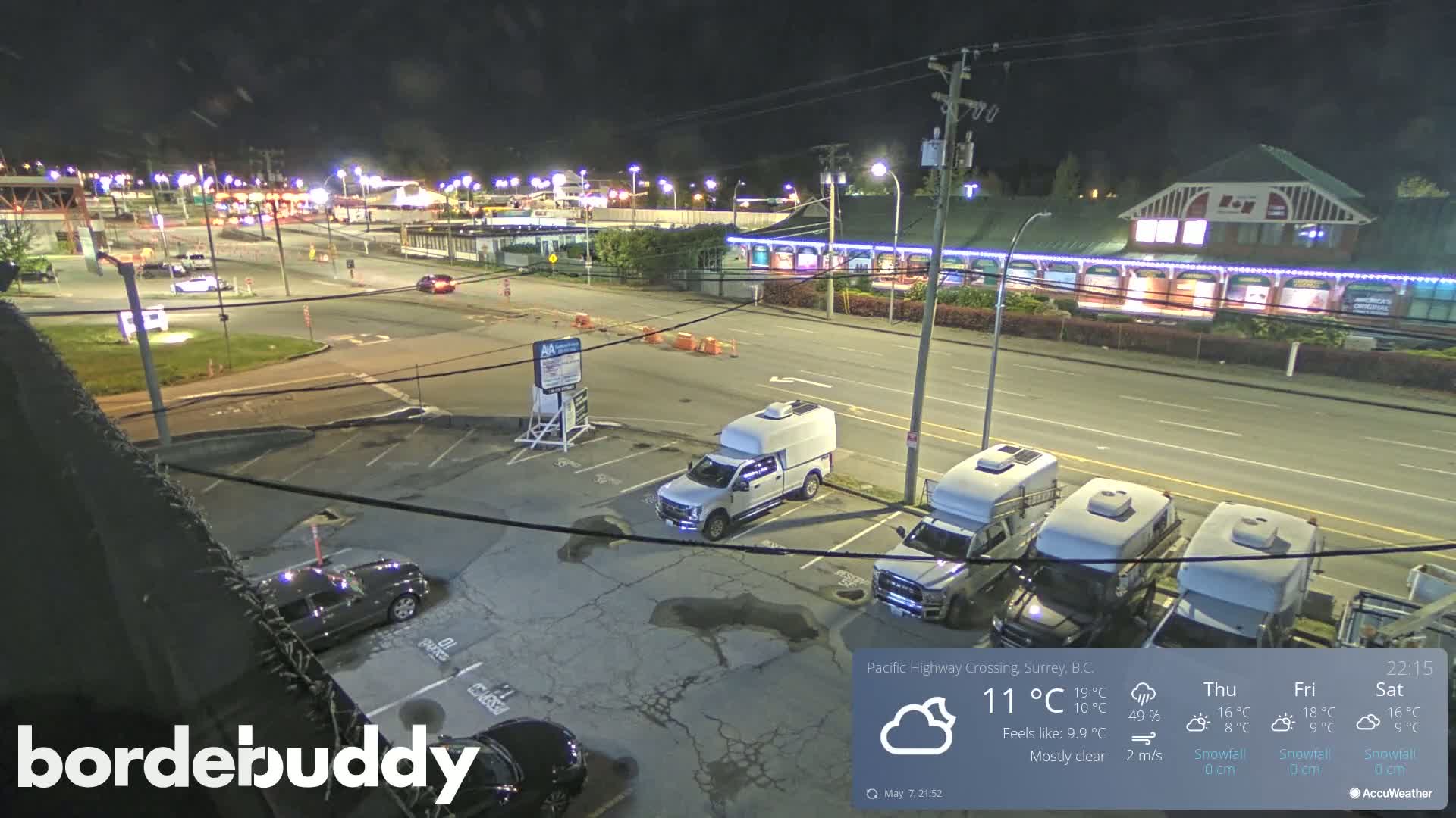 A mostly clear night scene shows a parking lot with several vehicles, including pickup trucks and RVs, near a road with some traffic and buildings under nighttime lighting.