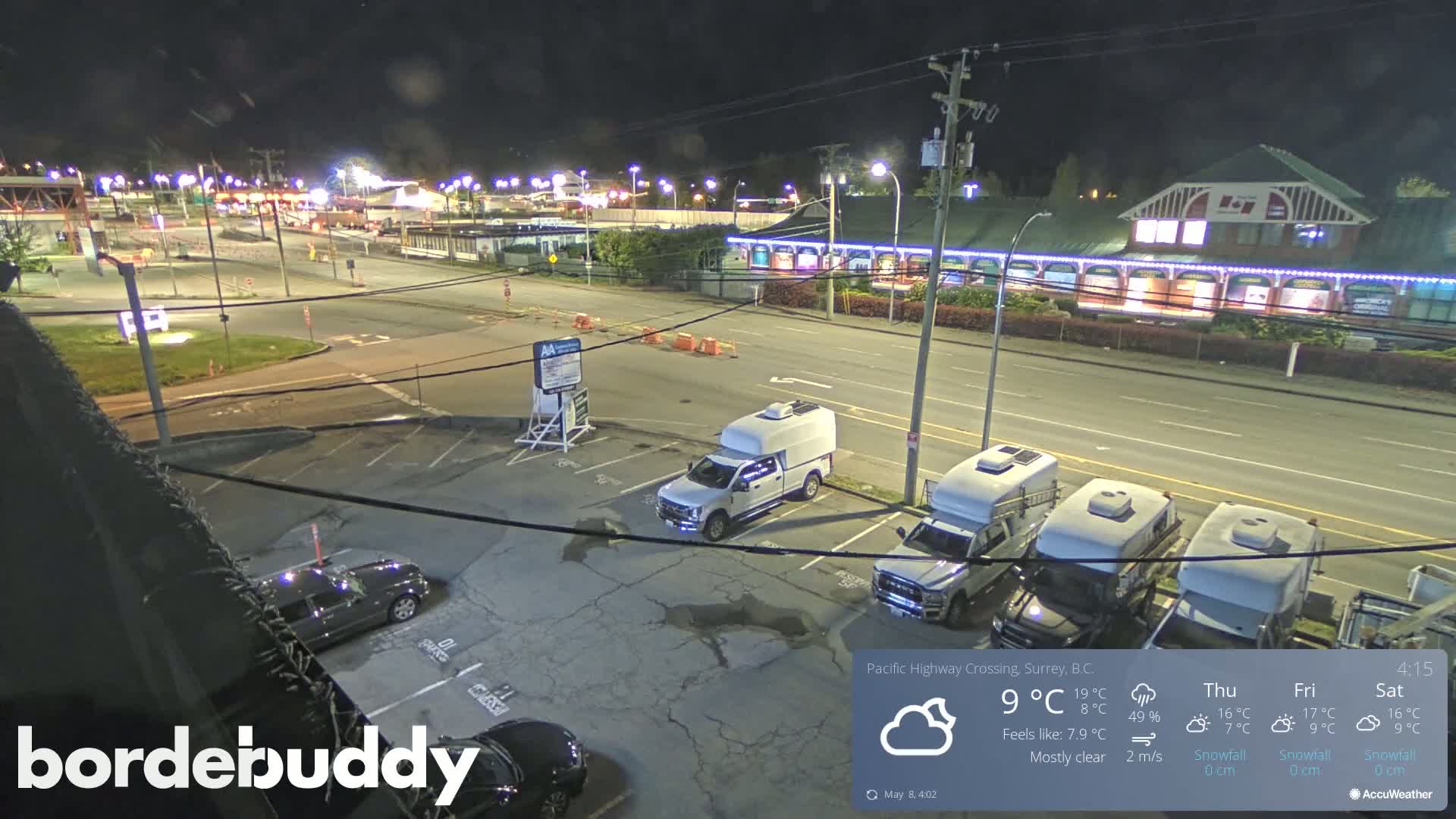 A nighttime view of a mostly empty parking lot next to a roadway, with several vehicles parked, under a mostly clear sky.