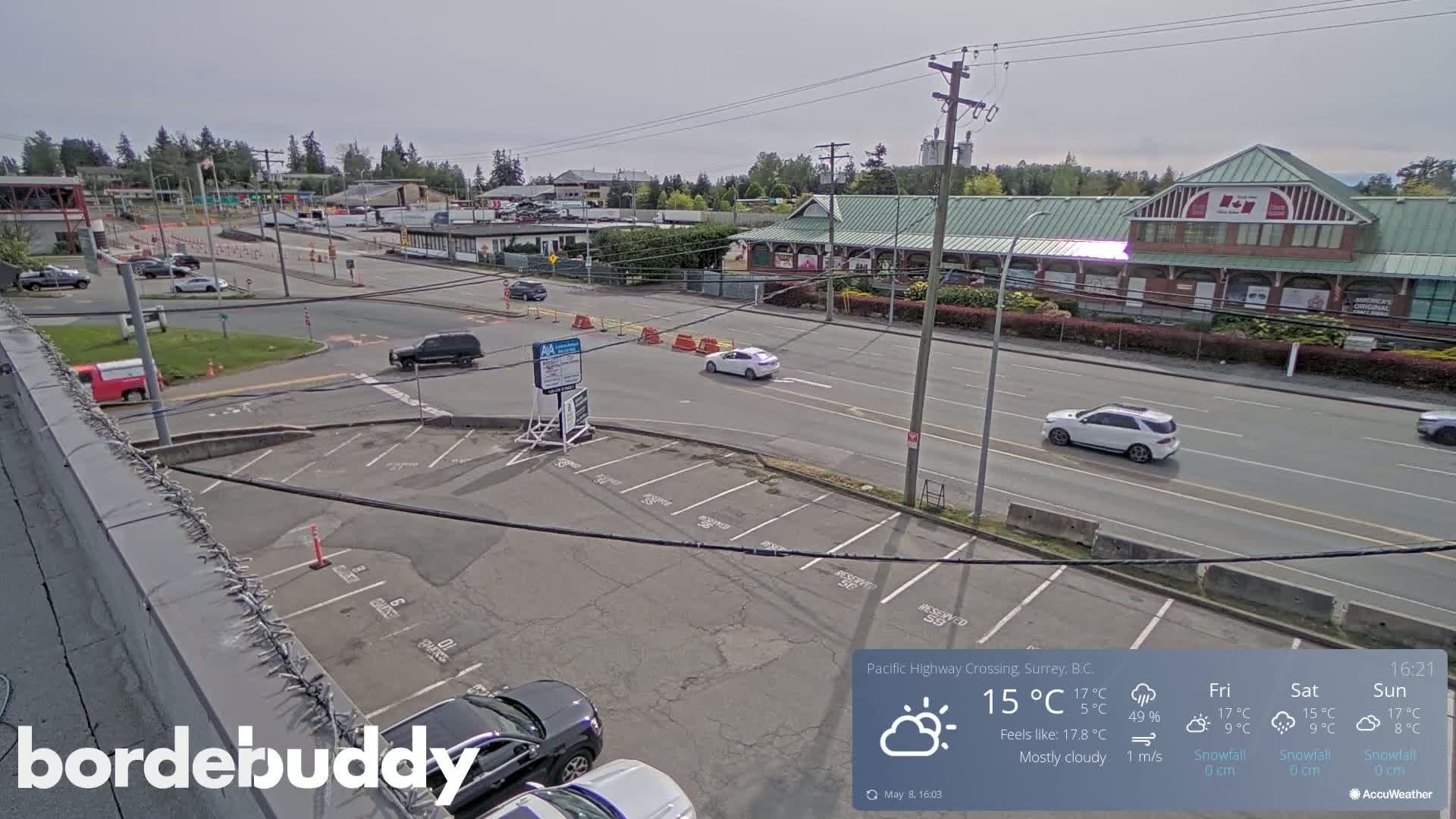 A mostly cloudy, overhead view shows a parking lot and a highway with several cars,  buildings, and some traffic control barriers.
