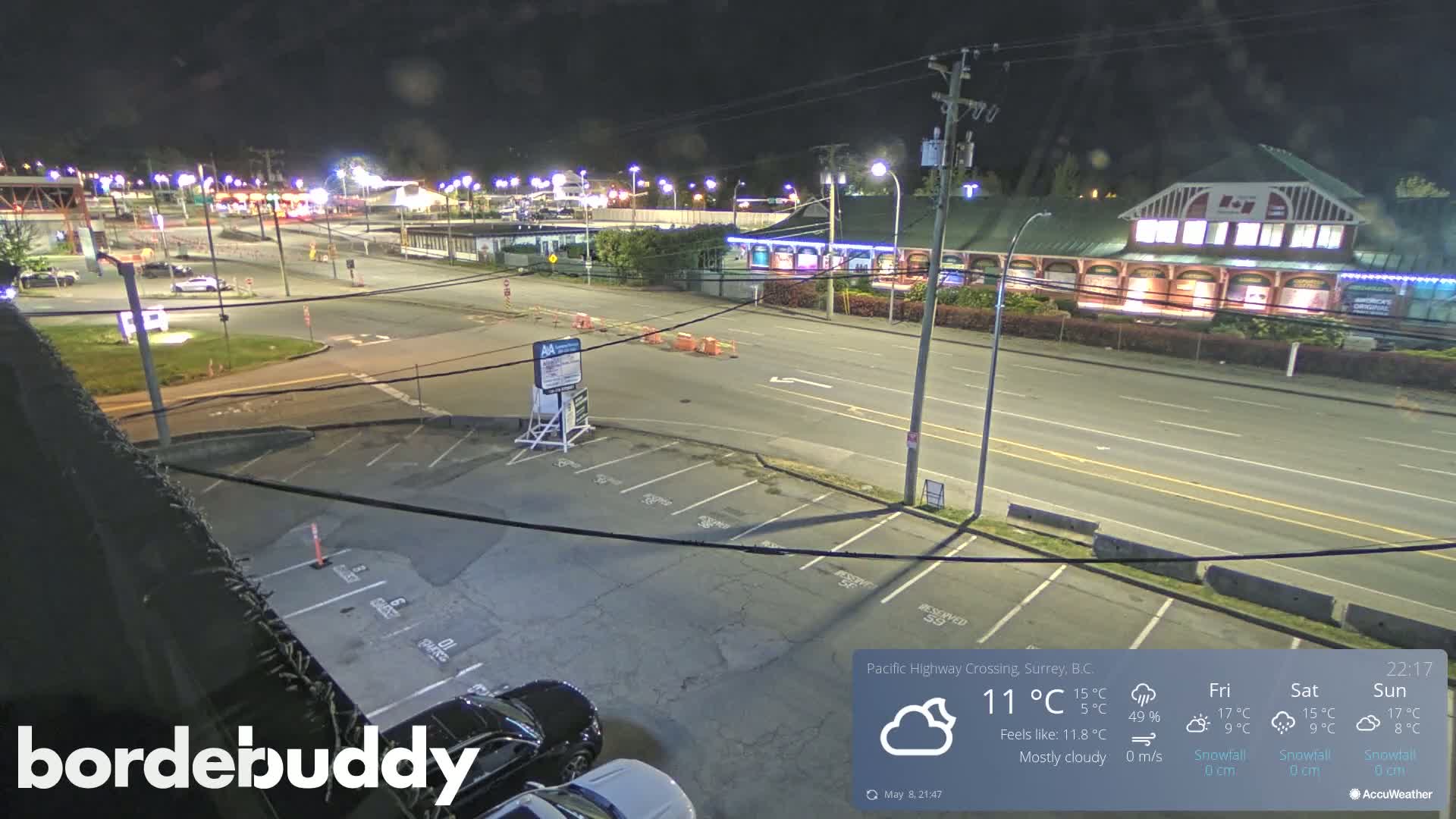 A mostly cloudy night scene shows a mostly empty parking lot next to a multi-lane road with some buildings and streetlights visible in the distance.